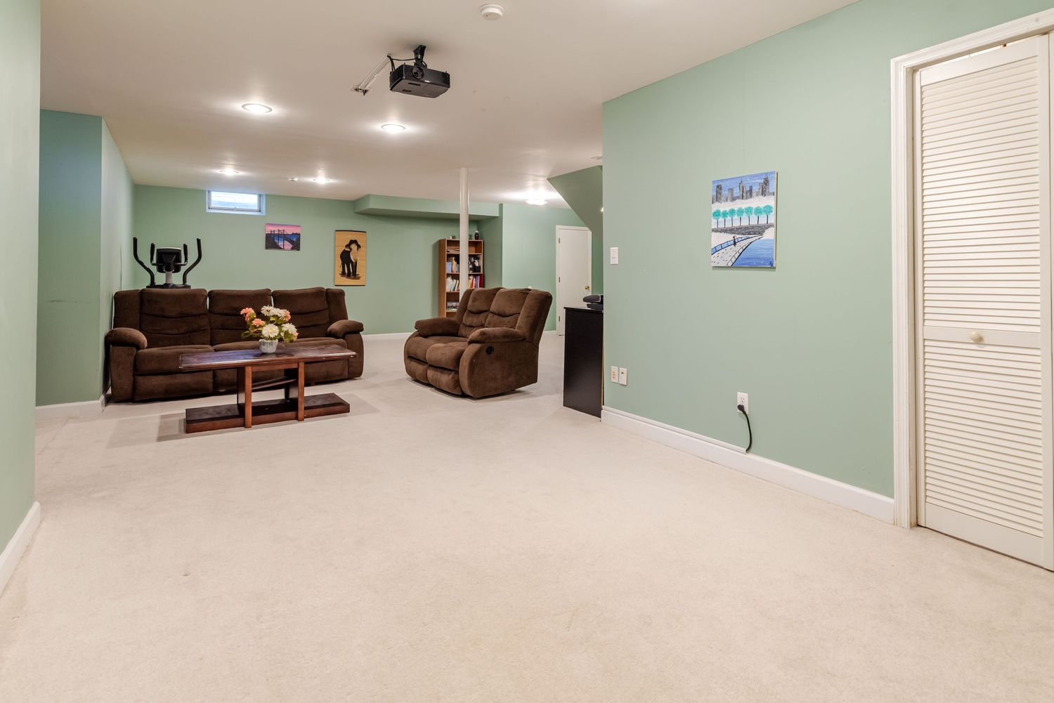 Spacious basement rec room with green walls, plush carpet, brown leather sofas, and a projector mounted on the ceiling.