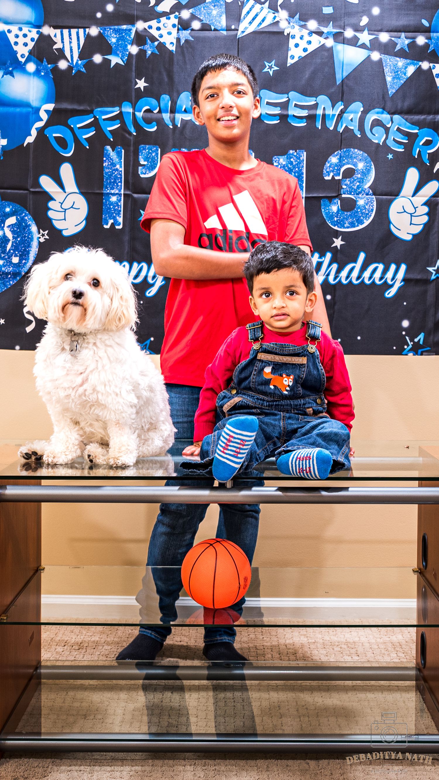 A small white dog and people sit on wooden steps against a blue birthday-themed backdrop with decorative text.
