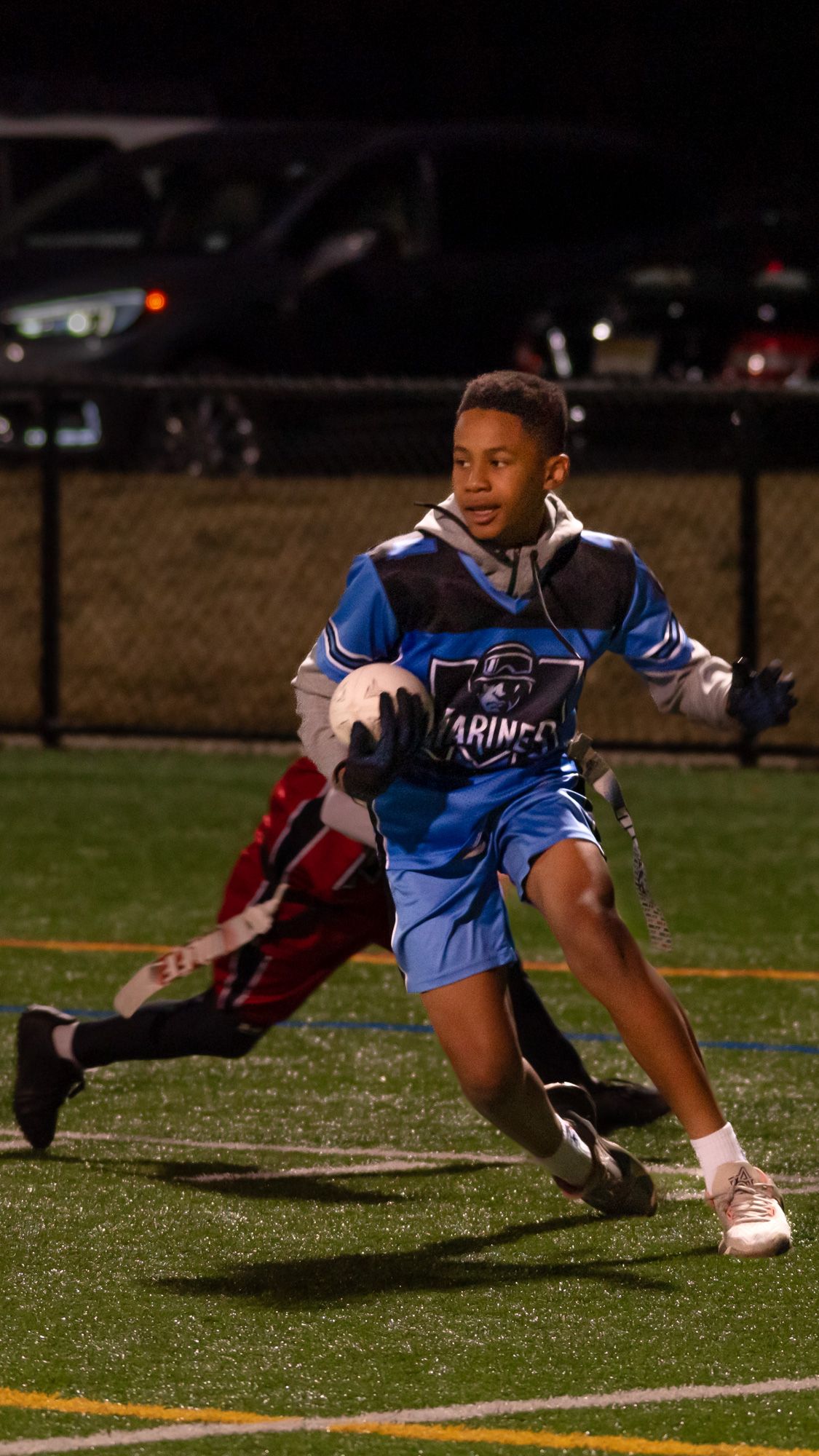 Young boy in blue Marines jersey runs with football during a night flag football game on turf field.