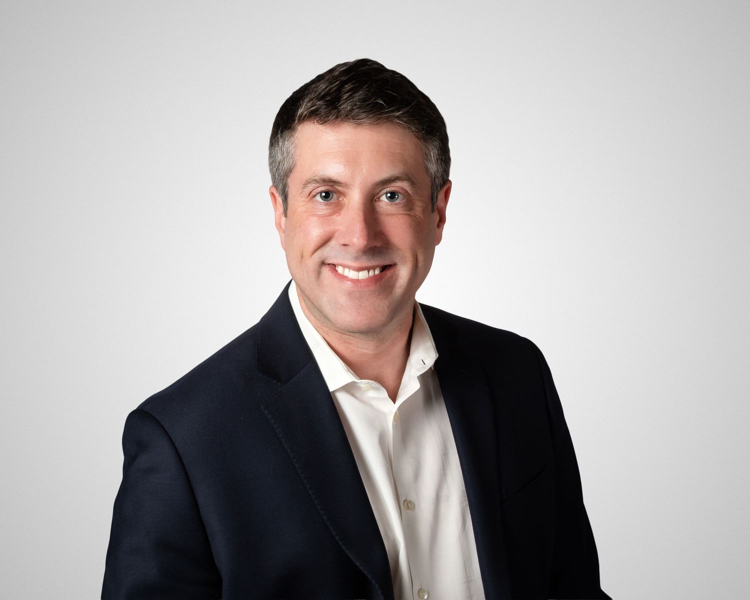 Professional headshot of a smiling middle-aged businessman in a navy blazer and white shirt against a gray background.