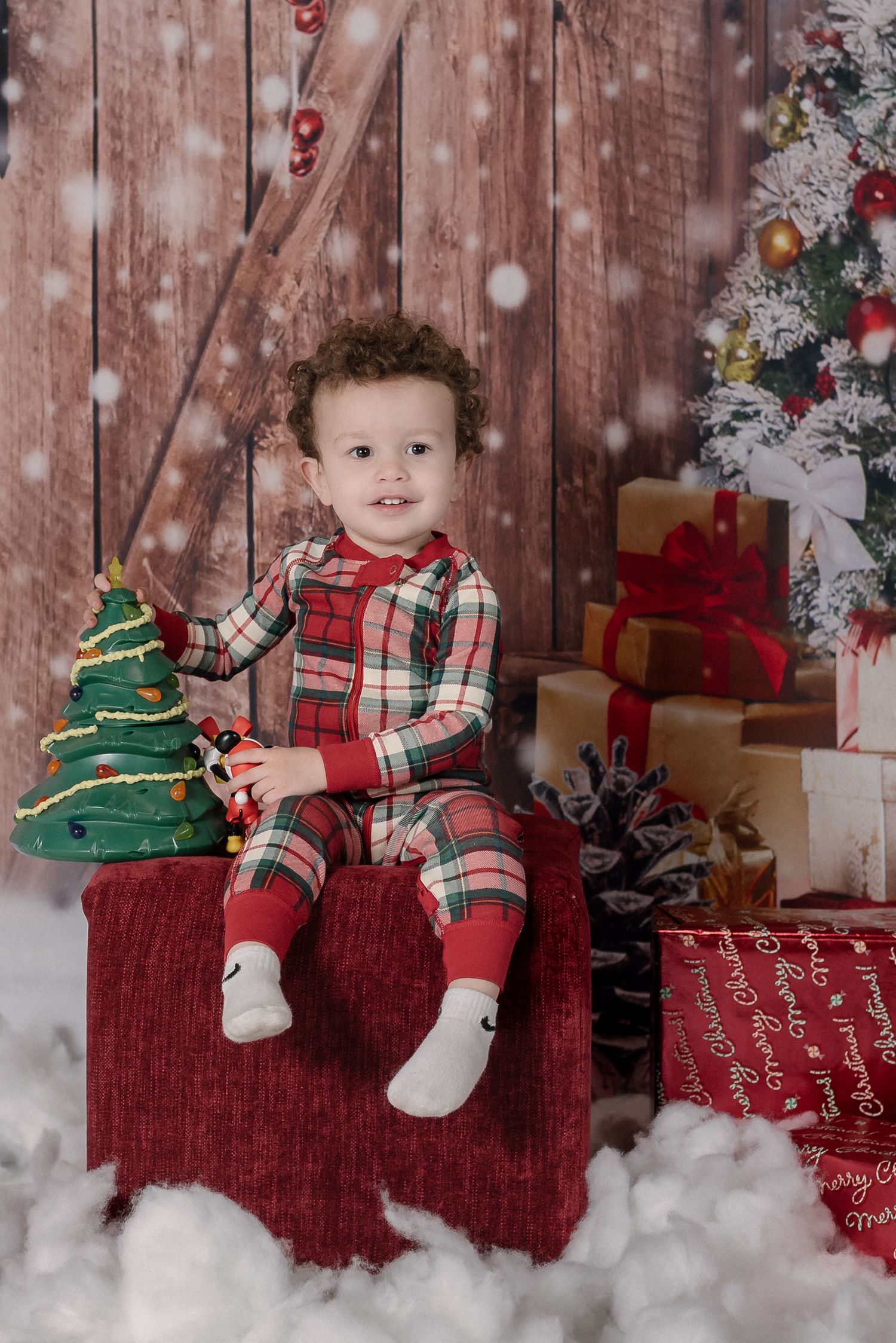 Christmas themed photo with child sitting on red bench near decorated tree and presents in snowy winter scene.