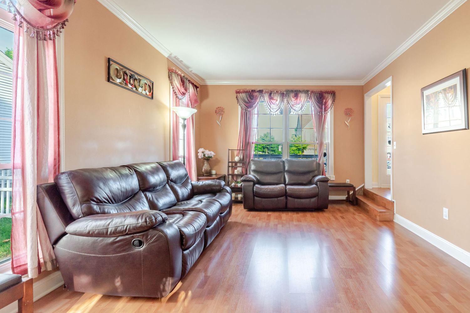 Bright living room with leather recliner sofas on hardwood floors, crown molding, and large windows with pink curtains.
