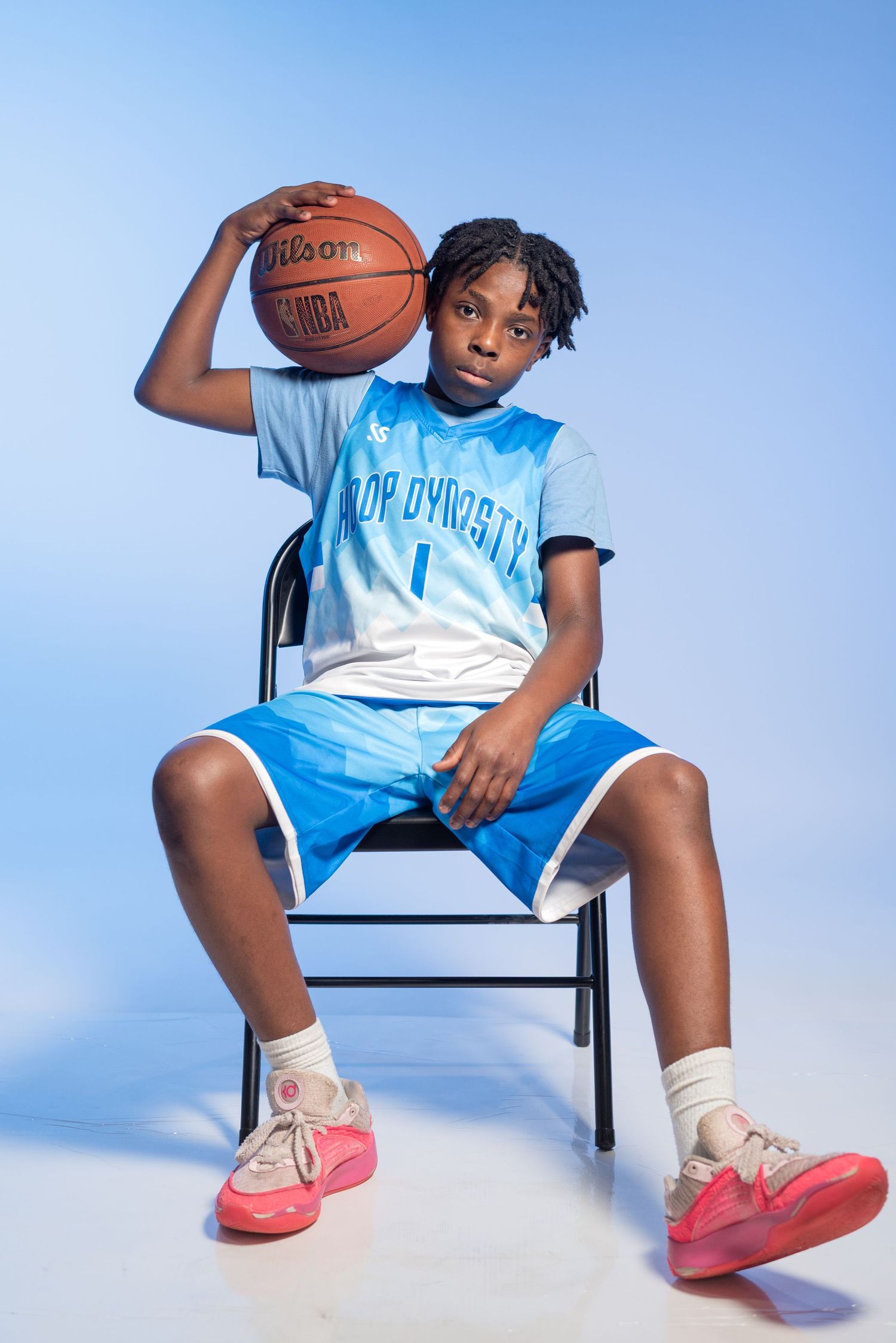 Young basketball player in blue Crop Dynasty jersey holding Wilson NBA ball while seated on chair against light blue background.