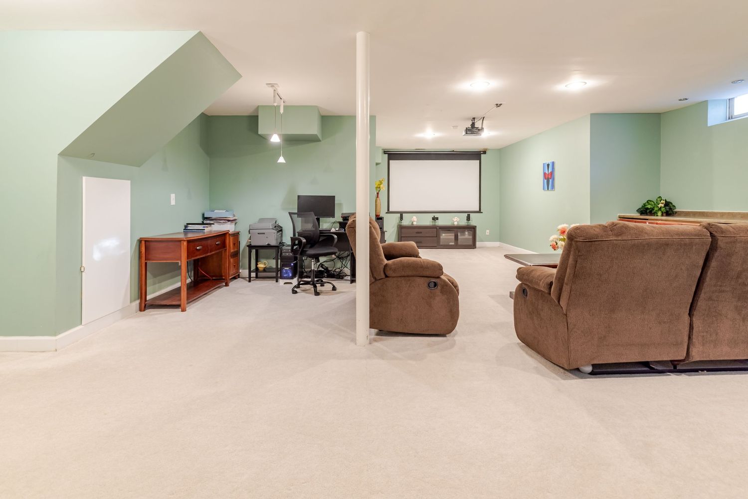 Spacious basement rec room with green walls, plush carpet, home office area, projection screen, and brown recliner sofa.