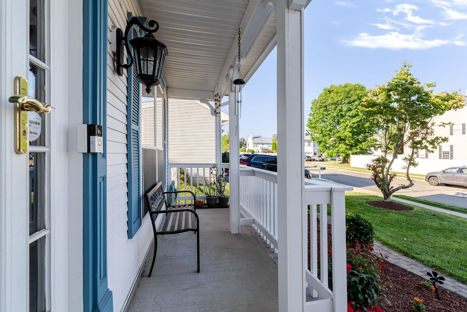 Charming front porch with black bench, blue door, lantern wall light, white railings, and lush green trees on a sunny day.