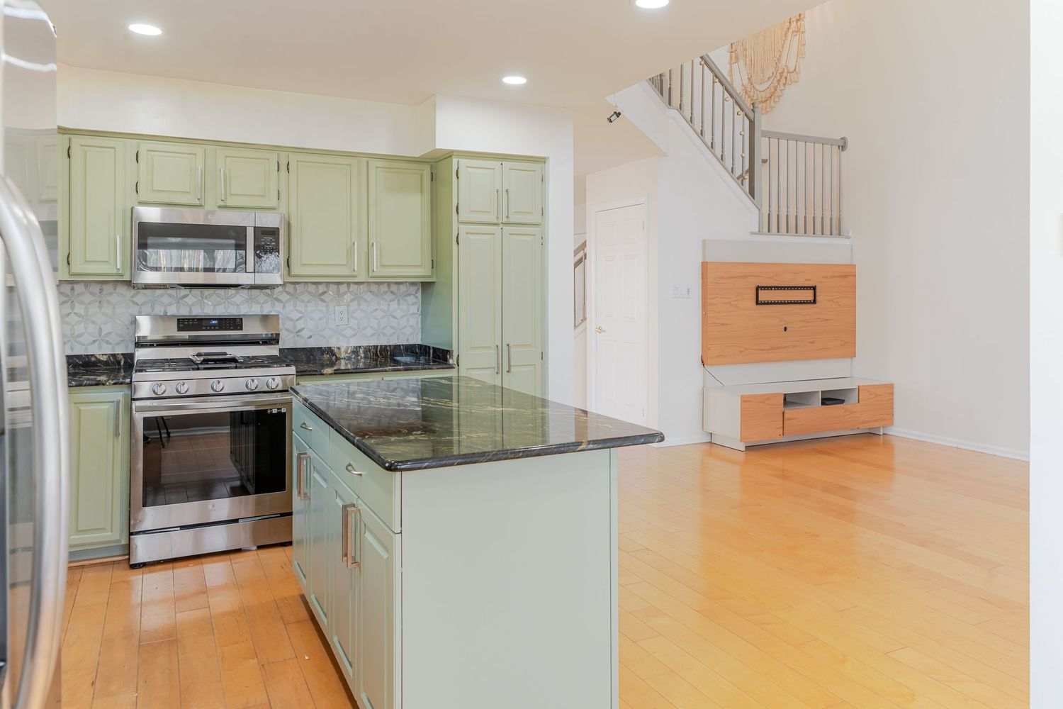 Modern kitchen with sage green cabinets, stainless steel appliances, dark granite island, and hardwood floors.