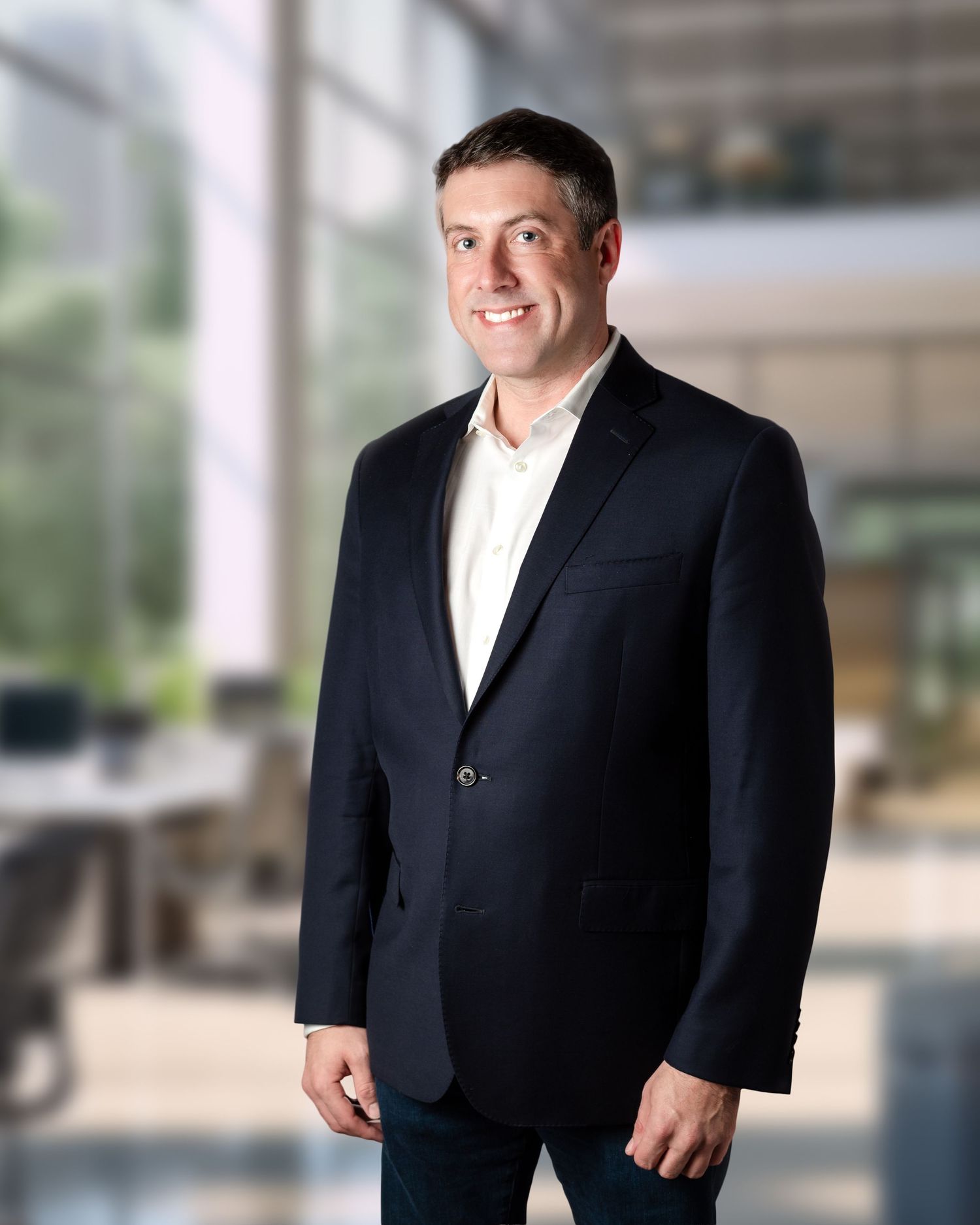 Professional businessman in dark blazer and white shirt smiling confidently in modern office setting.