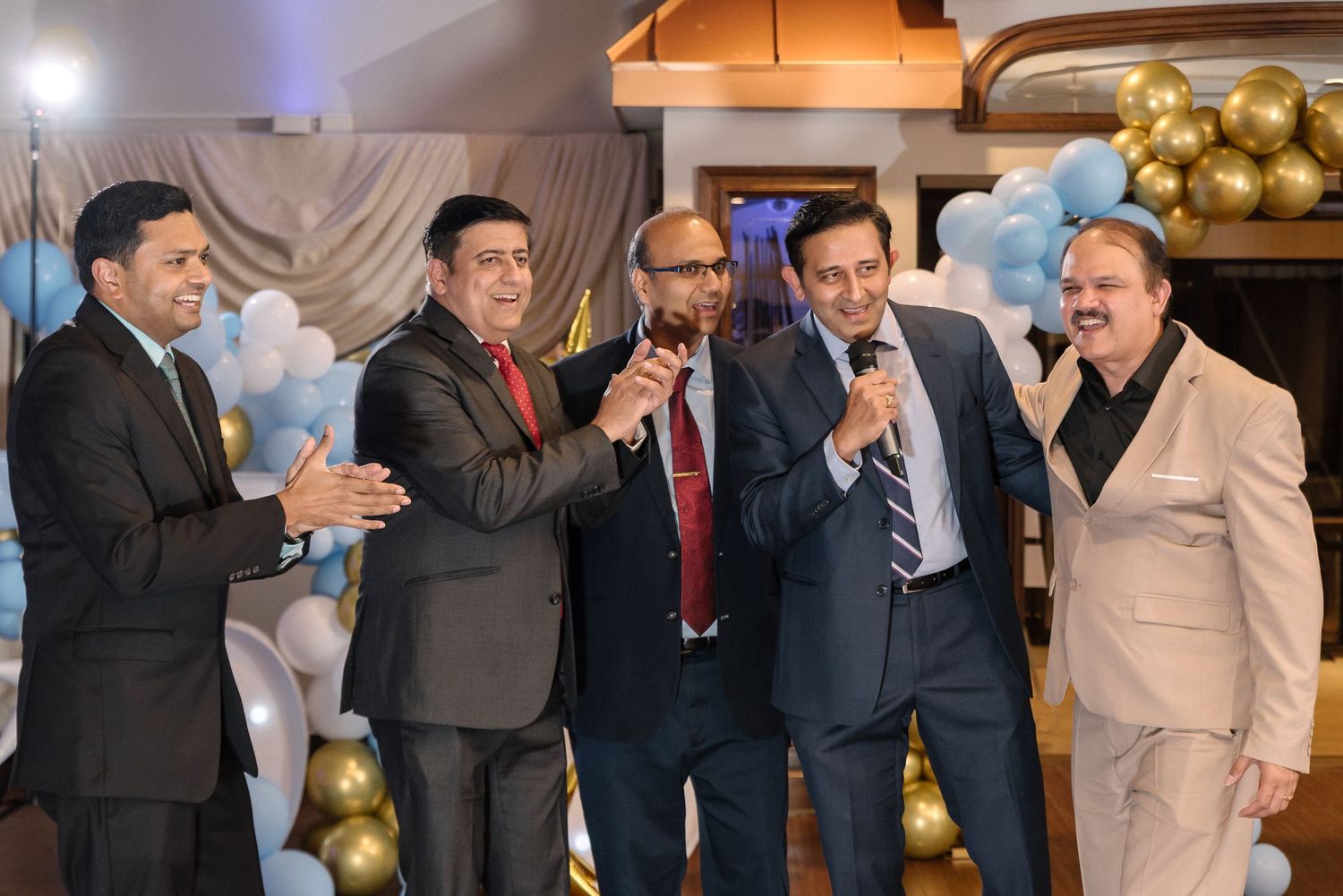 A group of people in formal attire celebrate at a social gathering with gold balloons and decorative lighting.