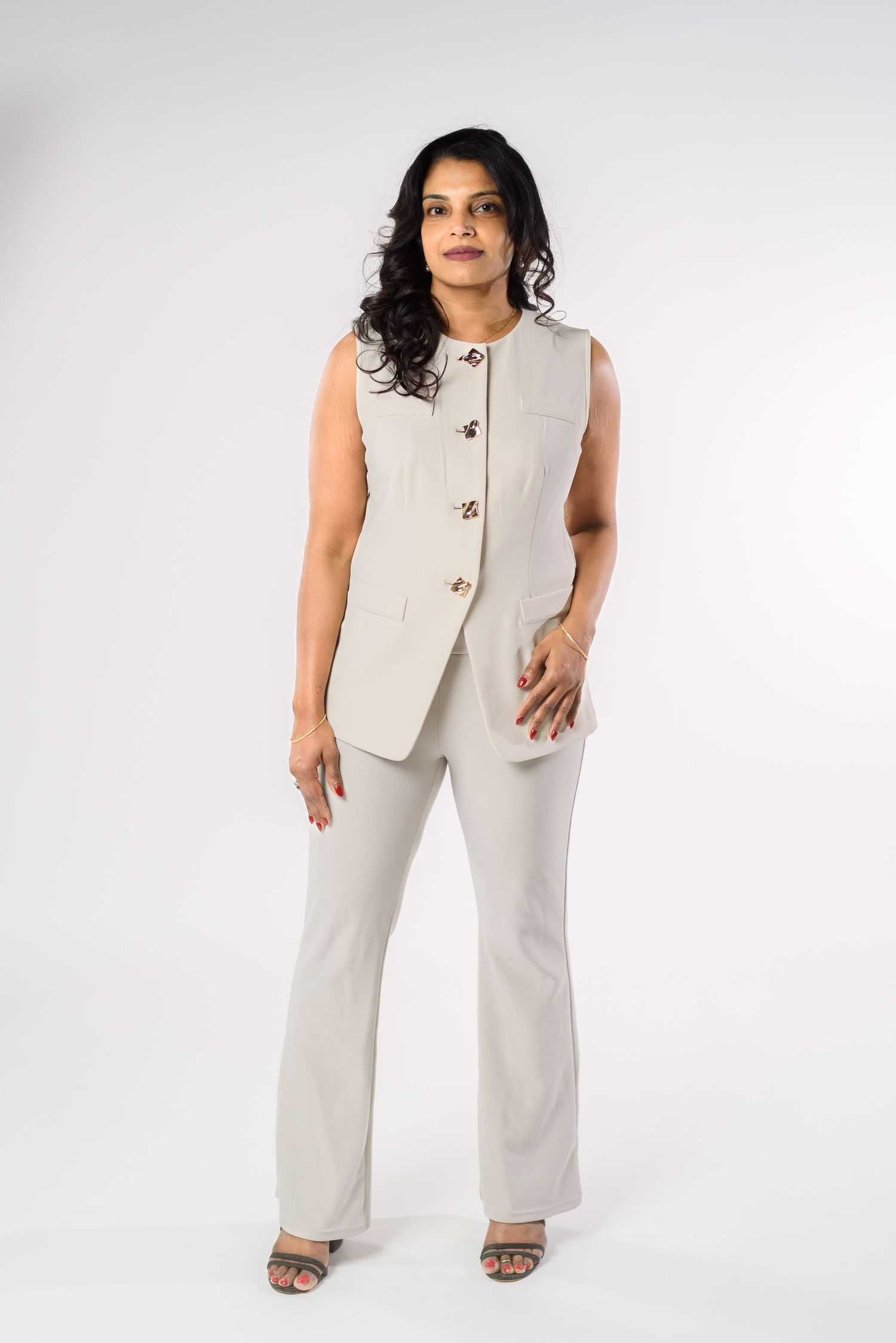Woman wearing a beige sleeveless vest and matching flared trousers with heeled sandals on white background.