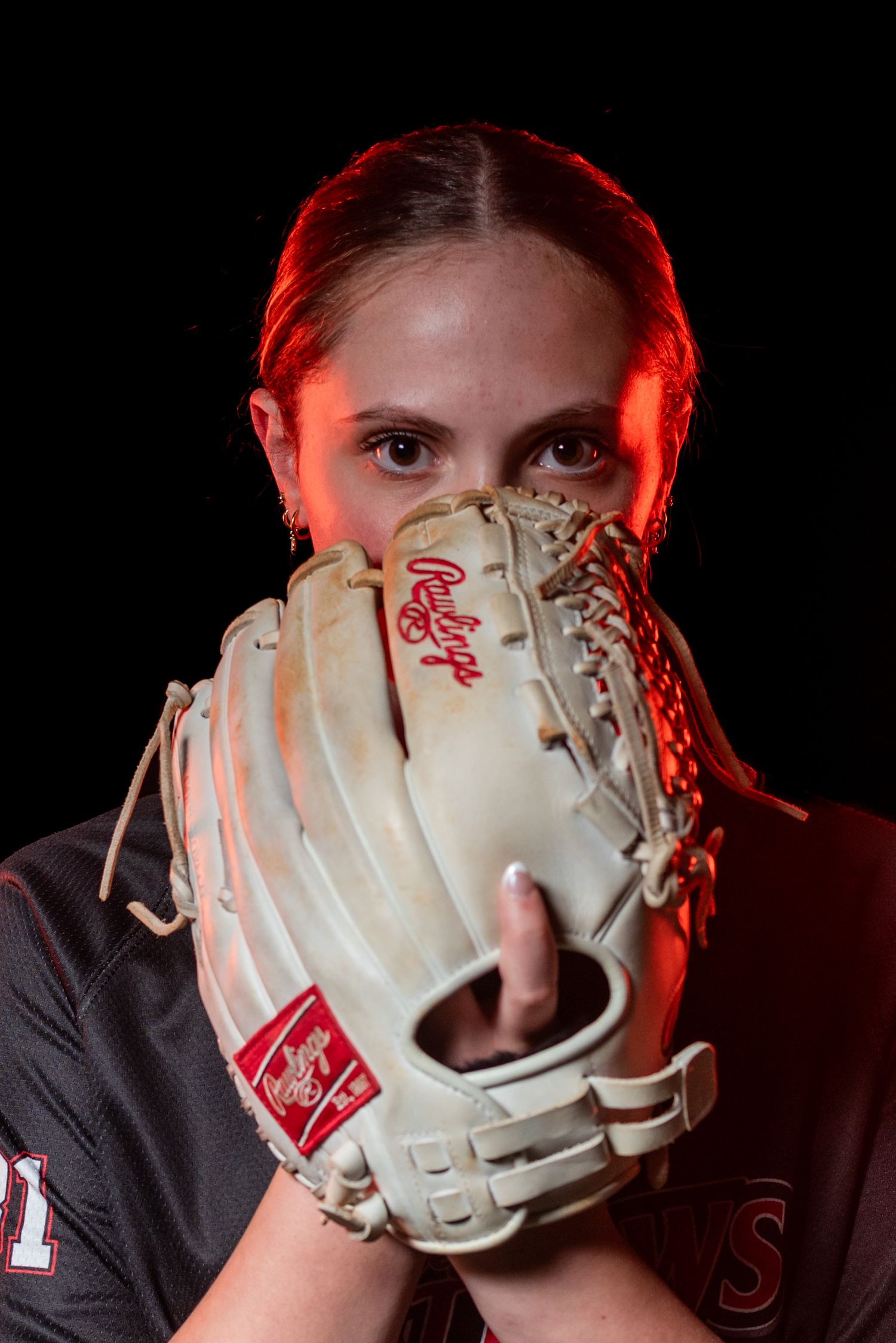 Female softball player holds a tan Rawlings glove up to her face, dramatically lit with red light against a black background.