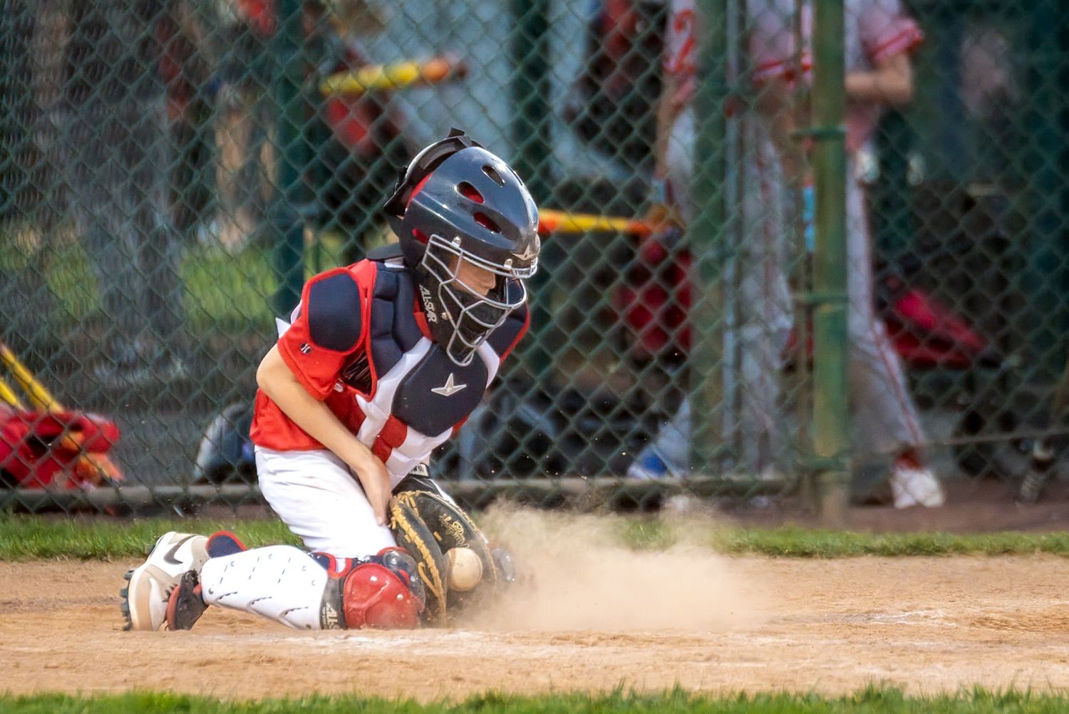 Youth baseball catcher in full gear blocking home plate in a cloud of dust during a little league game.
