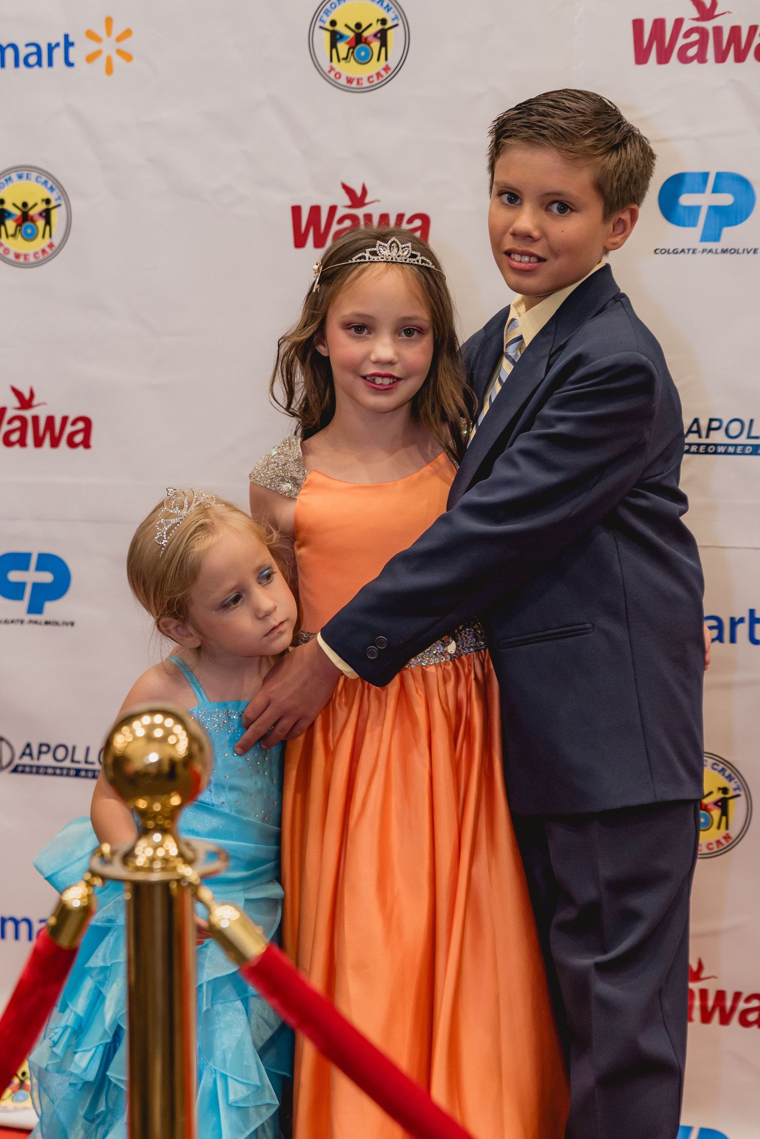 Children wearing orange and blue dresses pose at a formal event with branded backdrop.
