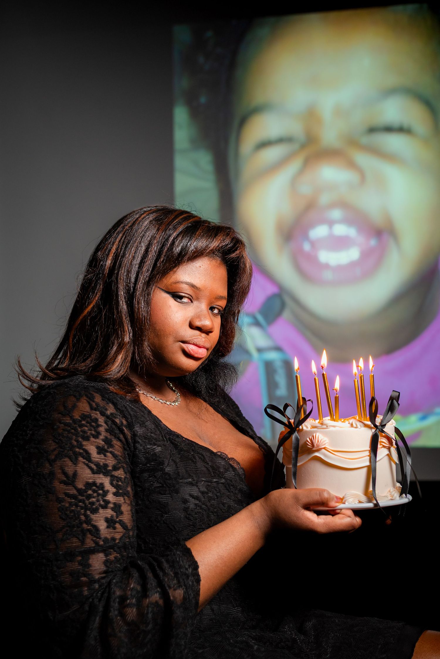 A young woman in black lace holds a birthday cake with lit candles, with a childhood photo projected behind her.