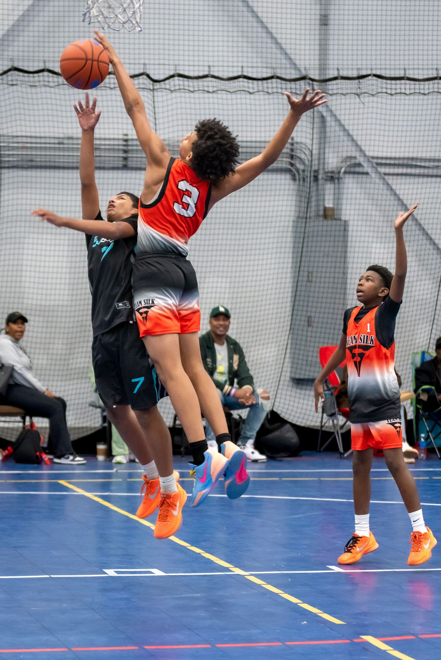 Youth basketball player in orange jersey number 3 leaps to block shot during indoor youth basketball game.