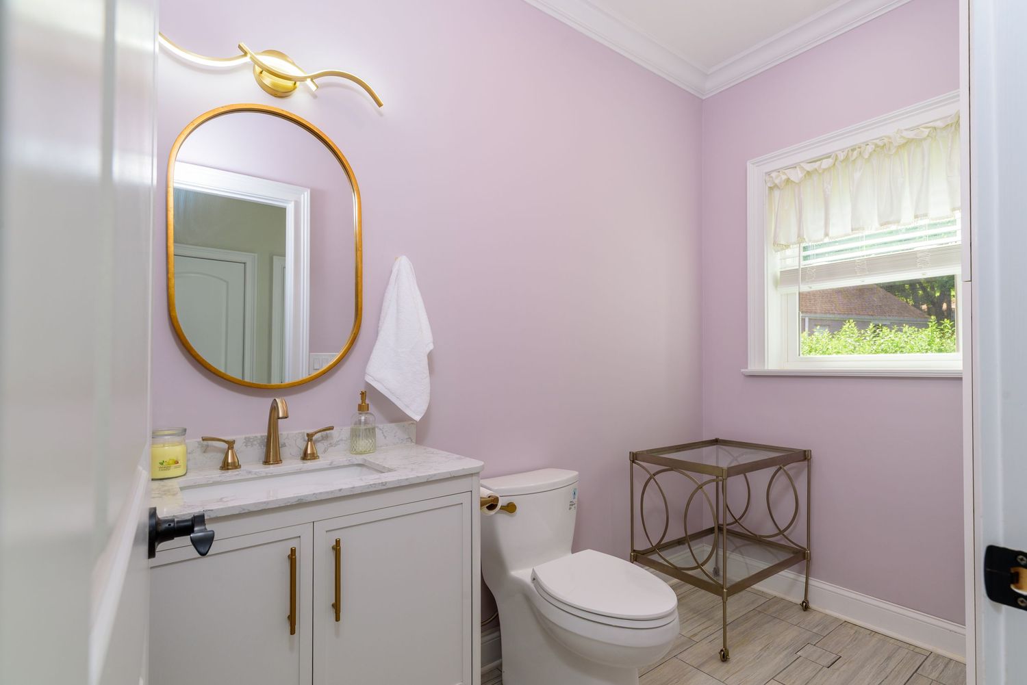 Pastel pink bathroom with gold accents, round mirror, and white fixtures.