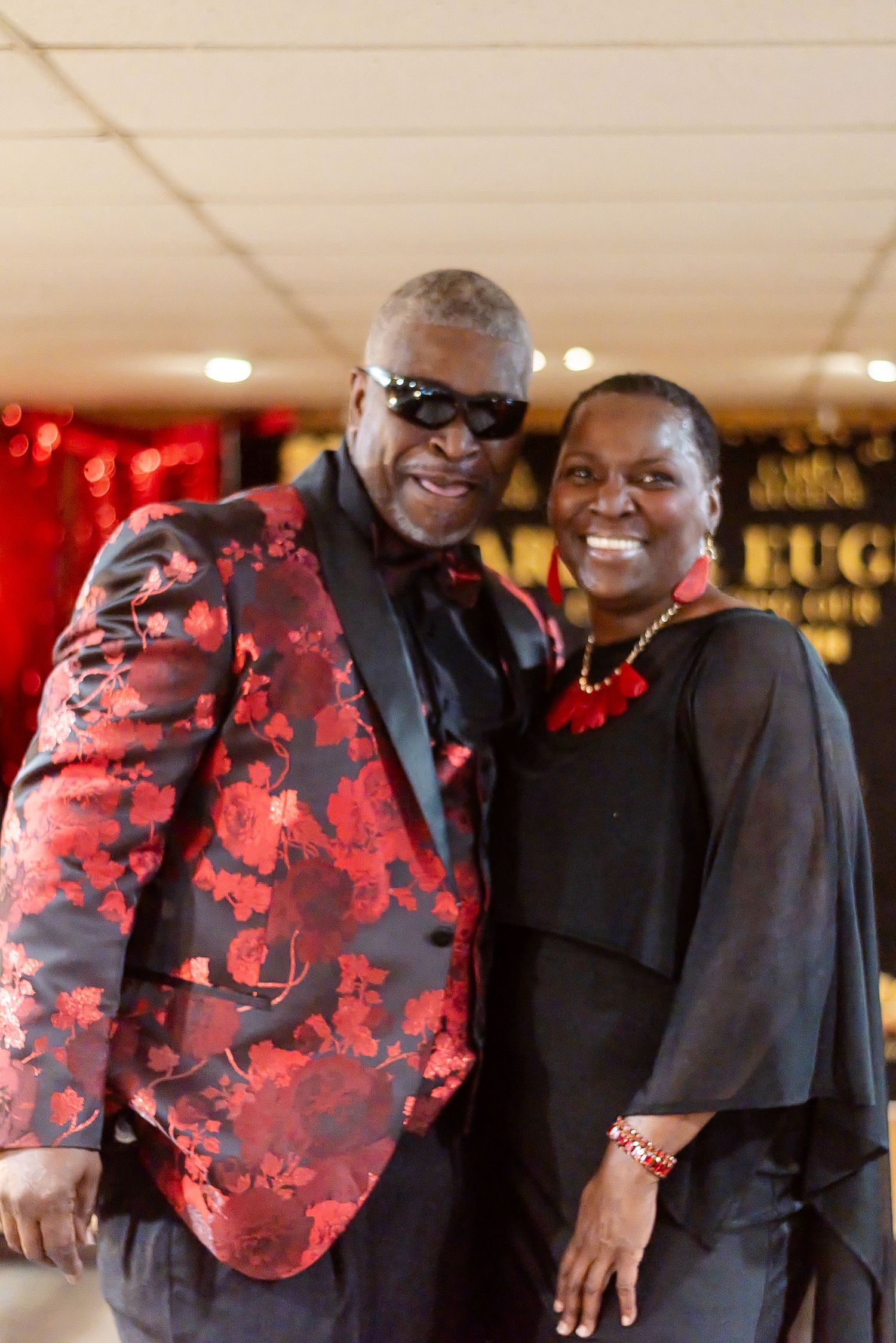 Couple posing together at formal event, man in red floral blazer with sunglasses, woman in black outfit with red accessories.