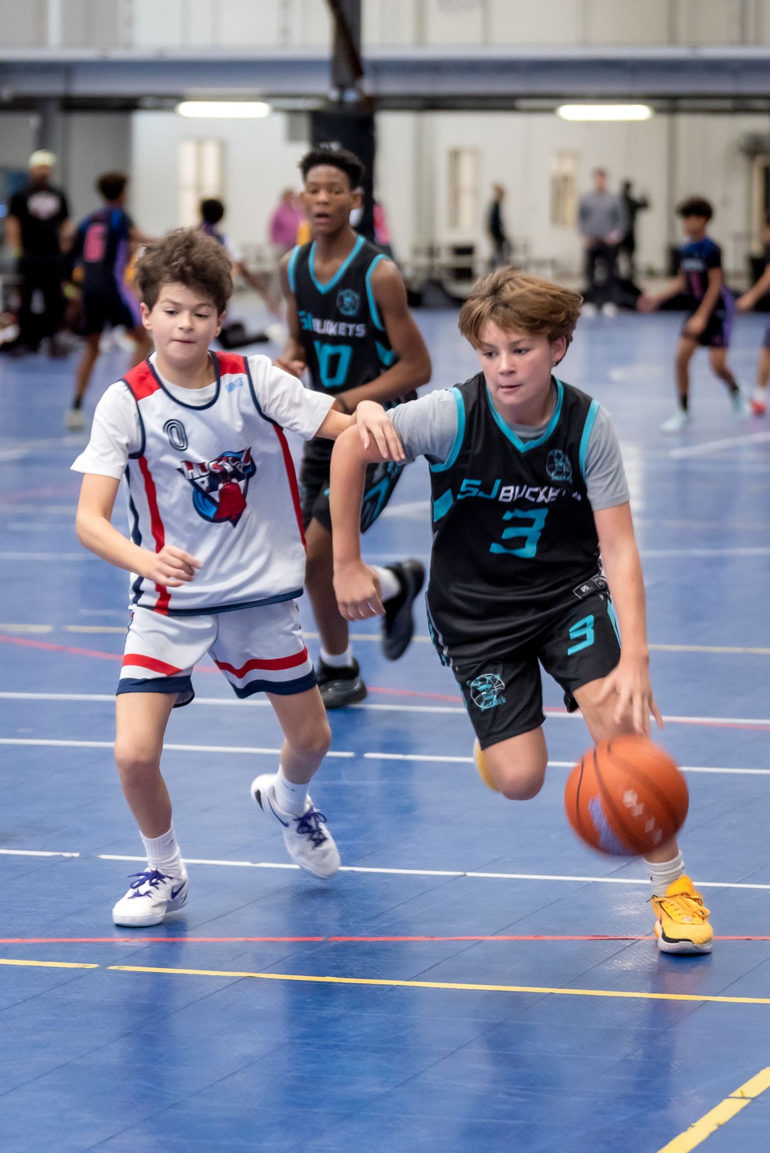 Two youth basketball players compete indoors, player in black jersey number 3 dribbling past defender in white uniform.