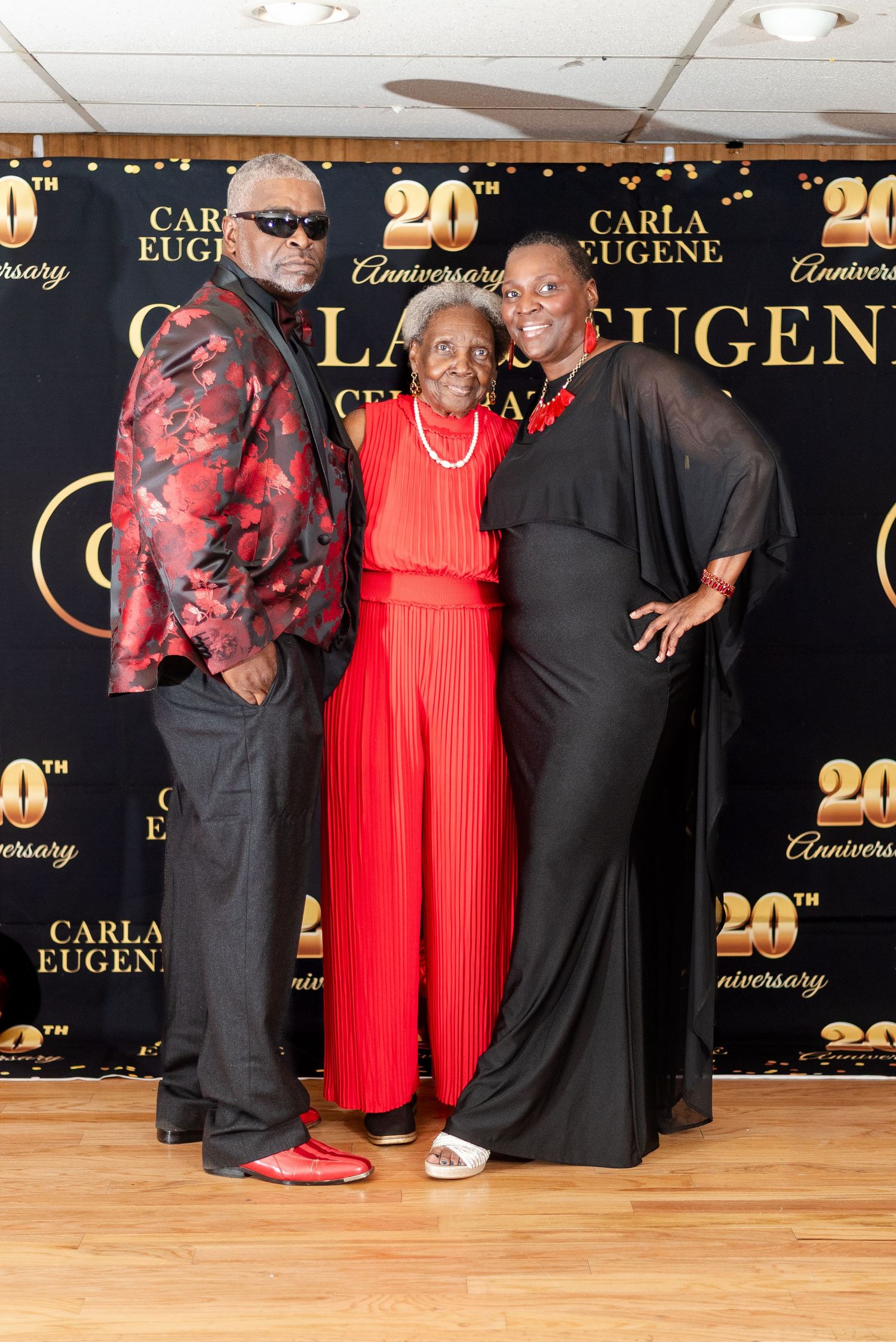 Three people pose at Carla Eugene's 20th Anniversary celebration, two in black outfits and one elderly woman in red.