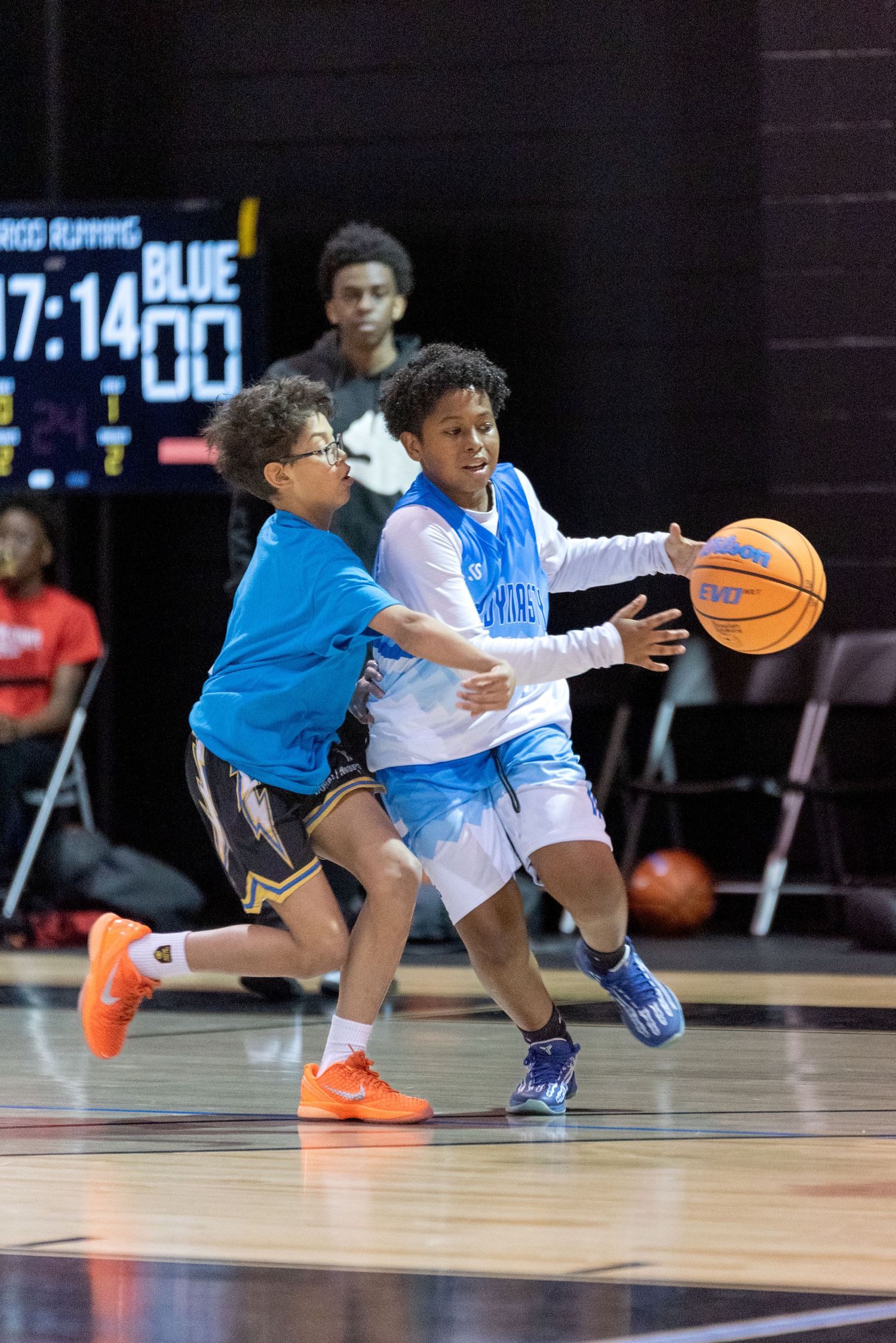 Two young boys playing basketball, one dribbling in a light blue jersey while the other defends in a teal shirt.