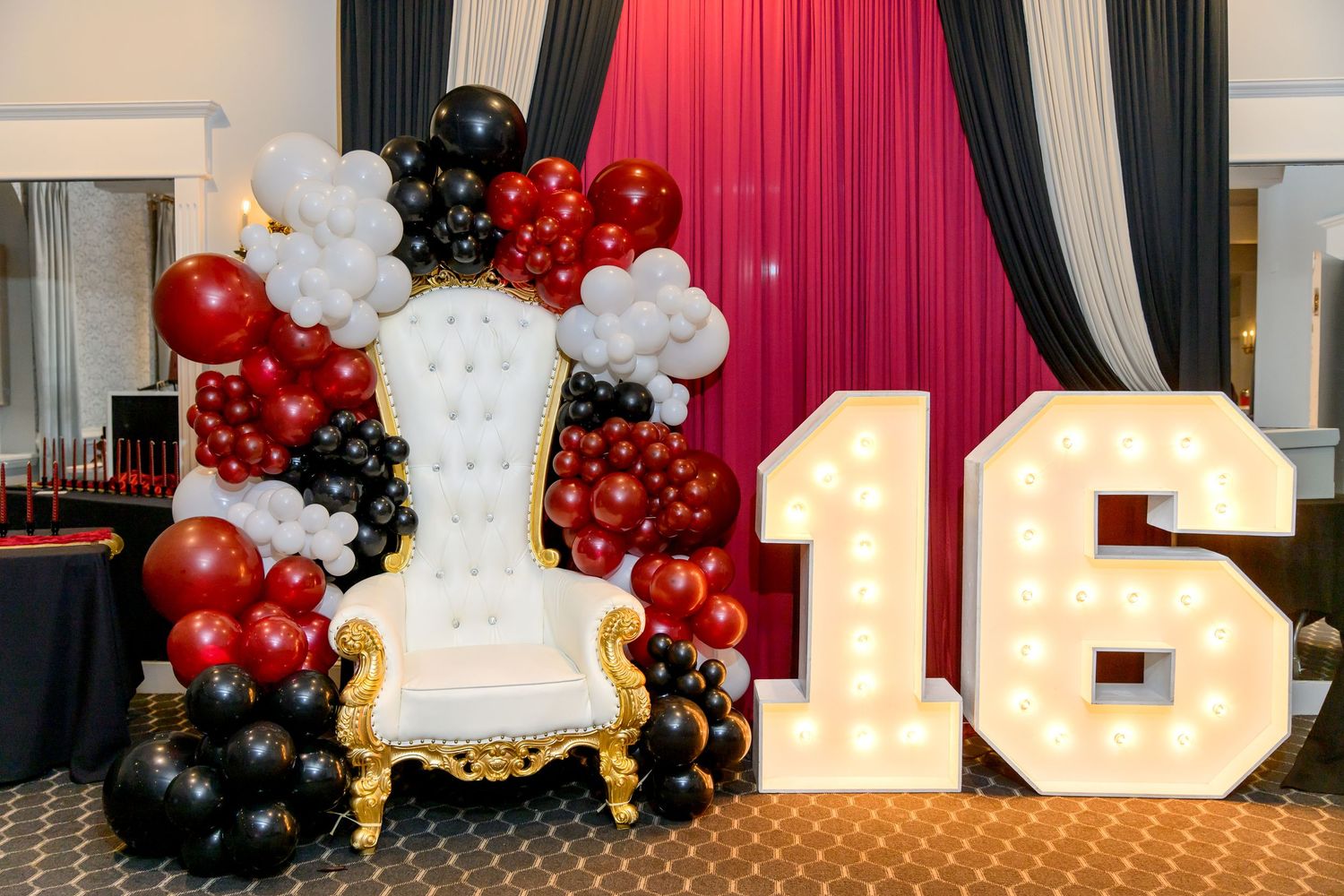 Sweet 16 party setup with white throne chair, marquee number 16, and red, black, white balloon garland.