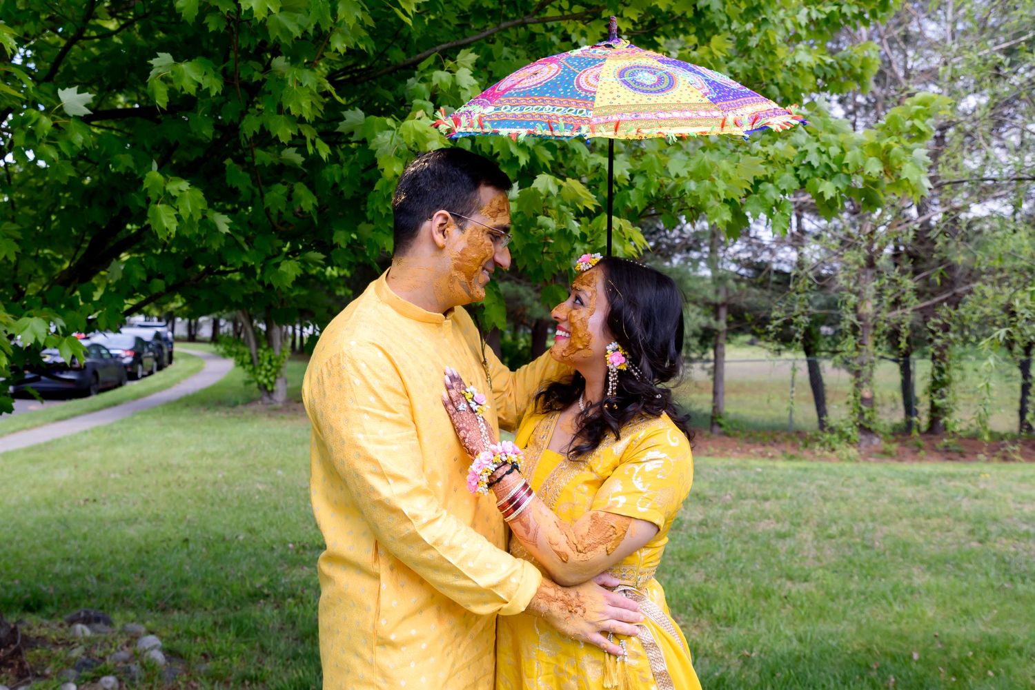 Couple in matching yellow outfits sharing a colorful umbrella outdoors, celebrating a traditional South Asian ceremony.