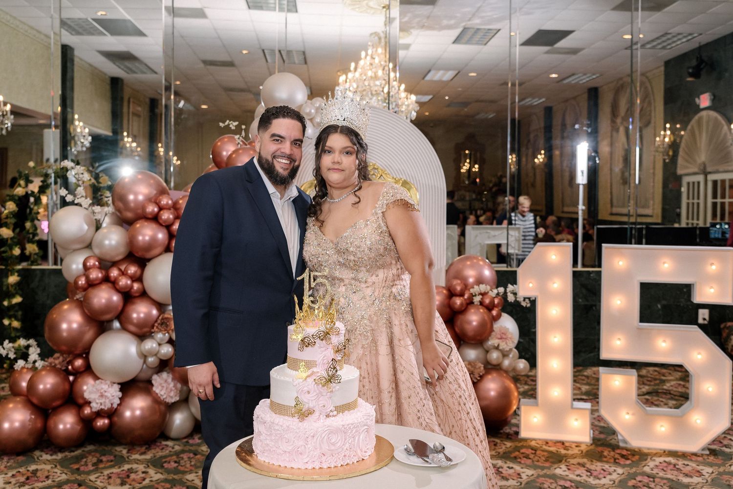 Couple posing by a quinceañera cake with balloon arch and marquee 15 sign at elegant venue.