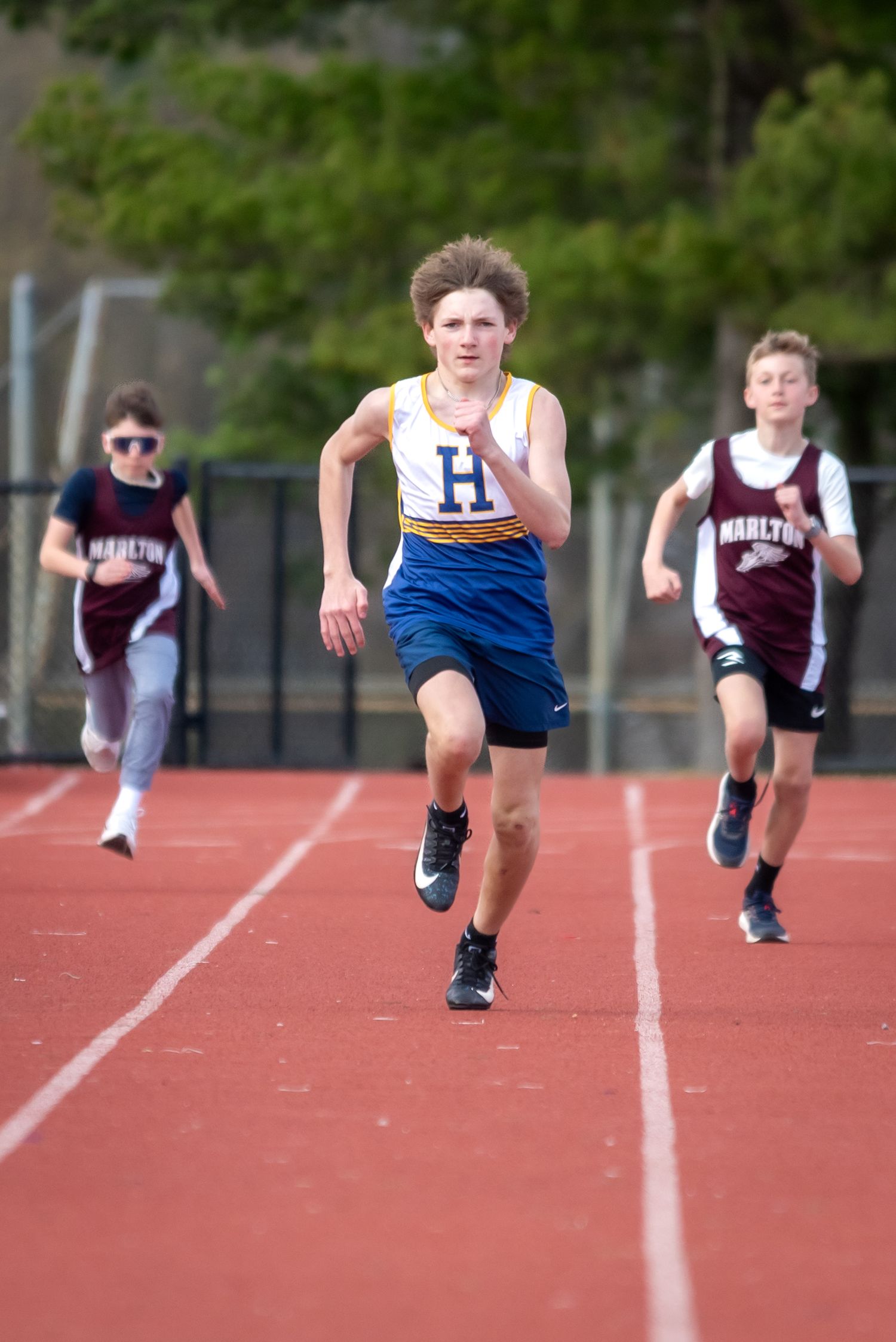 Young athlete in blue H jersey leads race on red track, outpacing competitors in maroon Marlton uniforms.