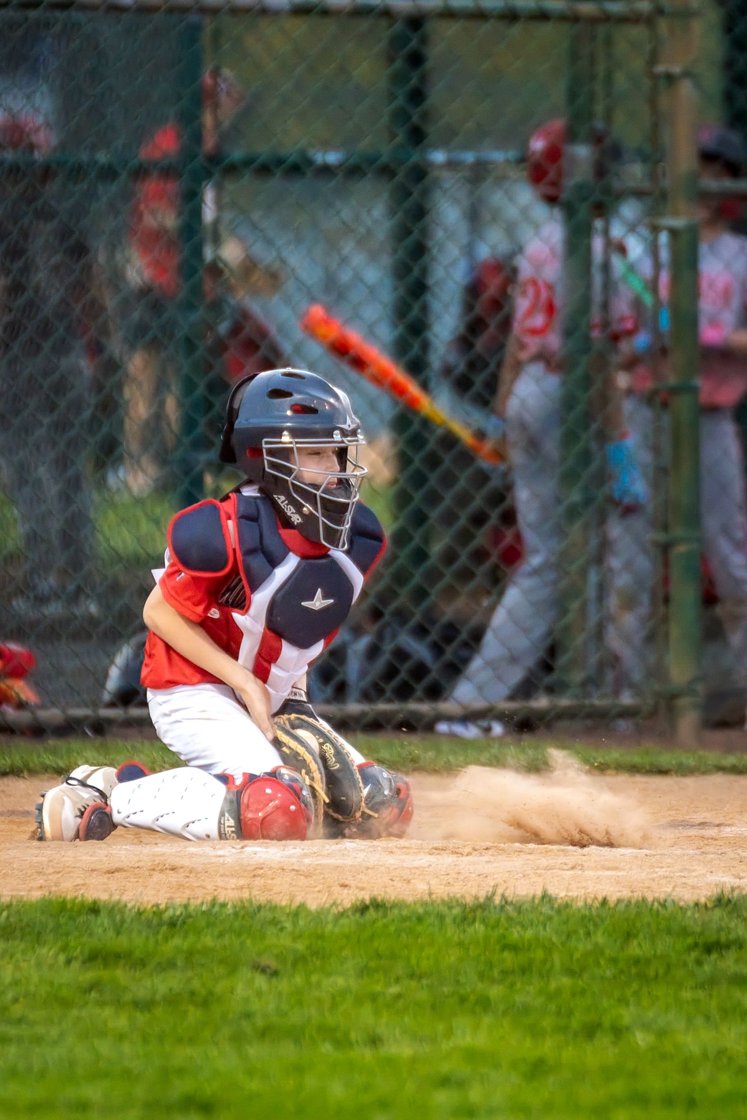 Young baseball catcher in red and navy uniform crouches at home plate during a youth league game.