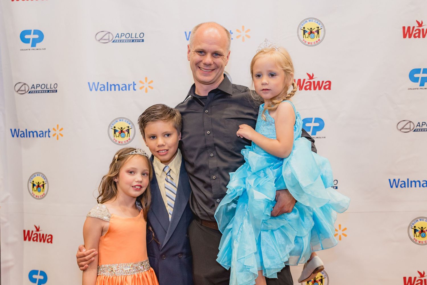 Children in formal attire pose at a red carpet event with sponsor logos in the background.