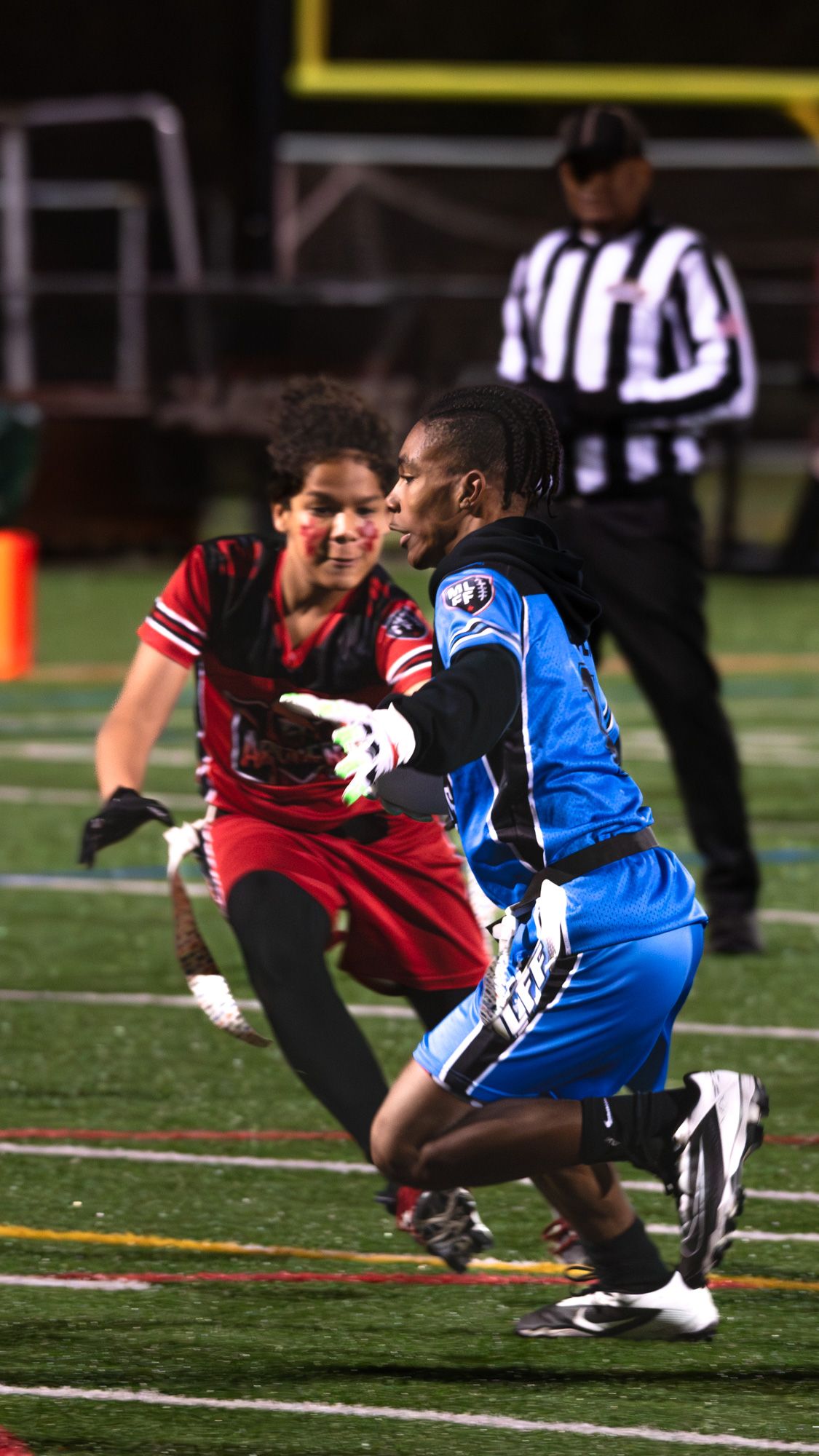 Two flag football players compete on a field at night, with a referee in black and white stripes visible in the background.