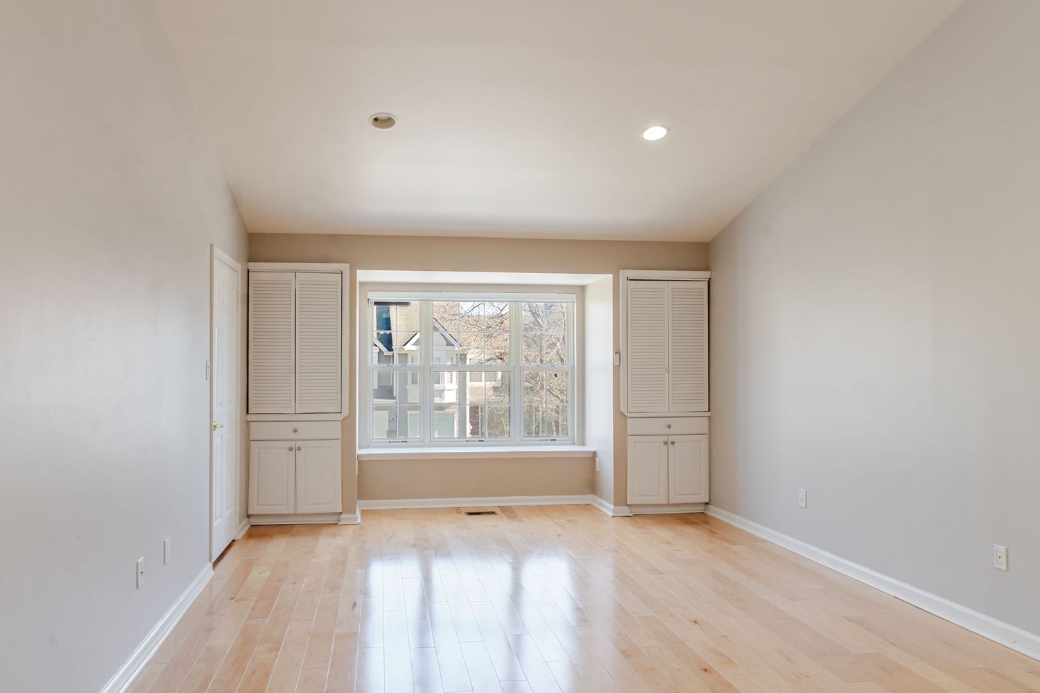 Bright empty room with built-in cabinets, large window, and gleaming hardwood floors.