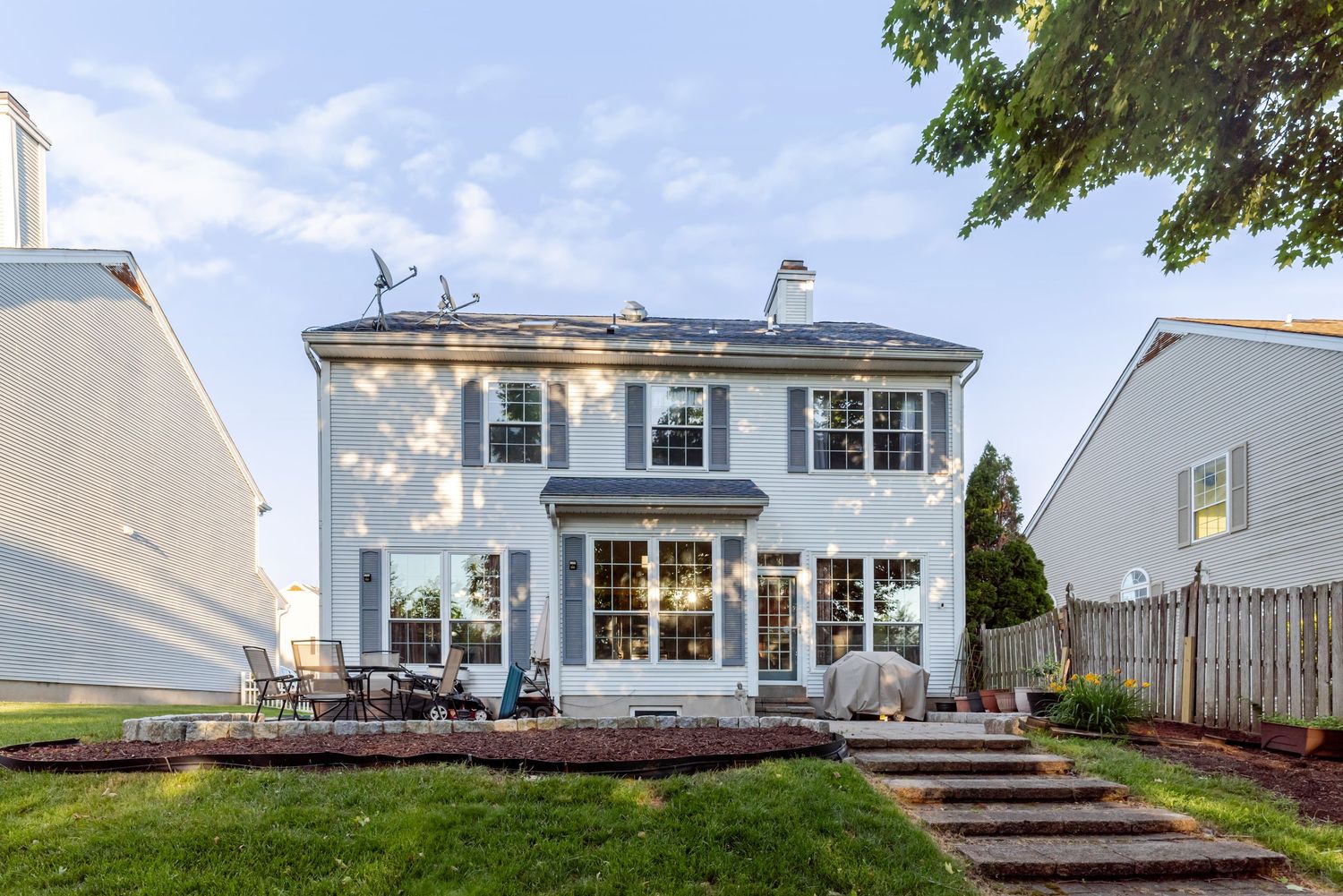White two-story colonial home with solar panels, patio seating, and lush green lawn in a suburban neighborhood.