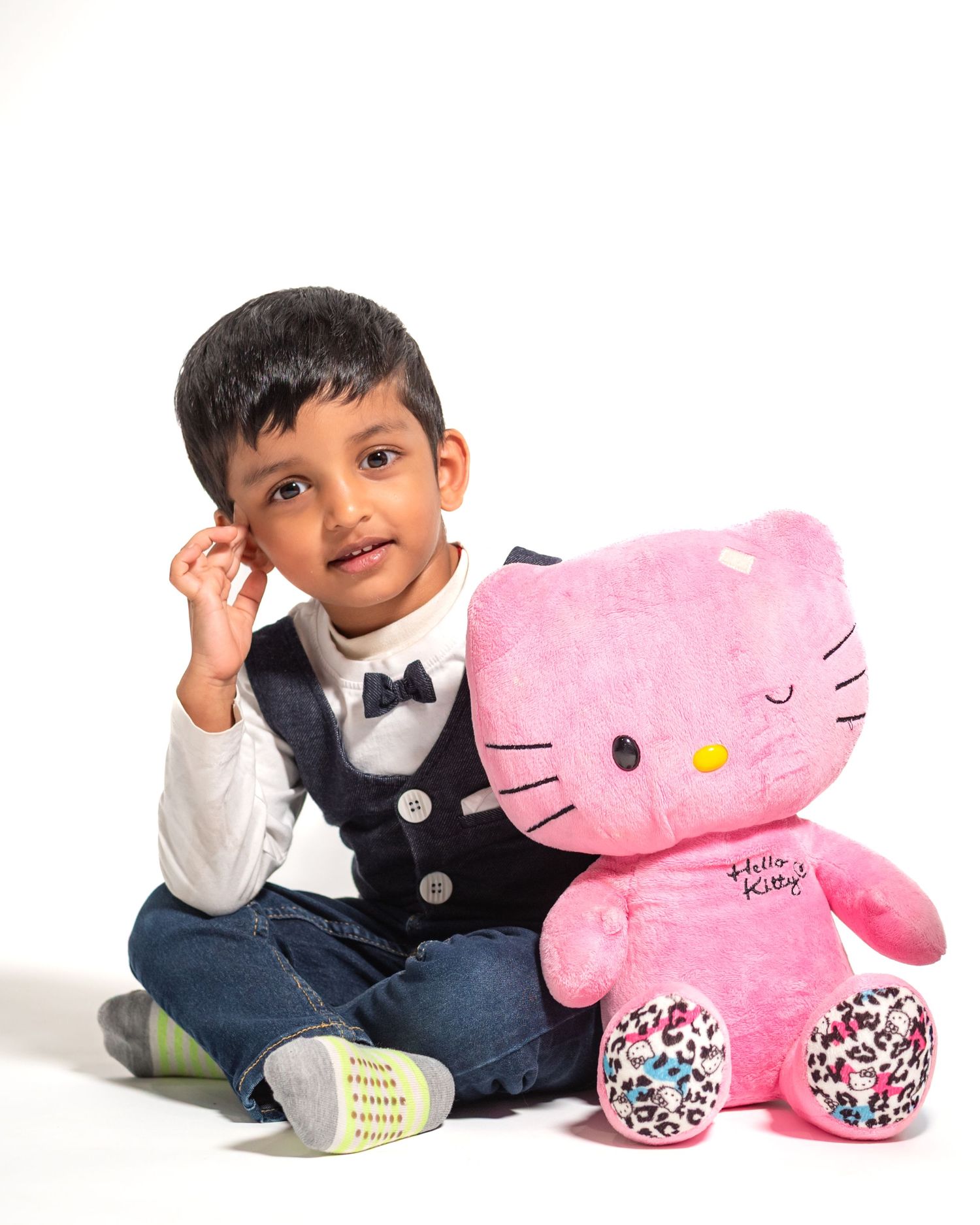 Young boy in vest and bow tie sits cross-legged beside a large pink Hello Kitty plush toy on white background.