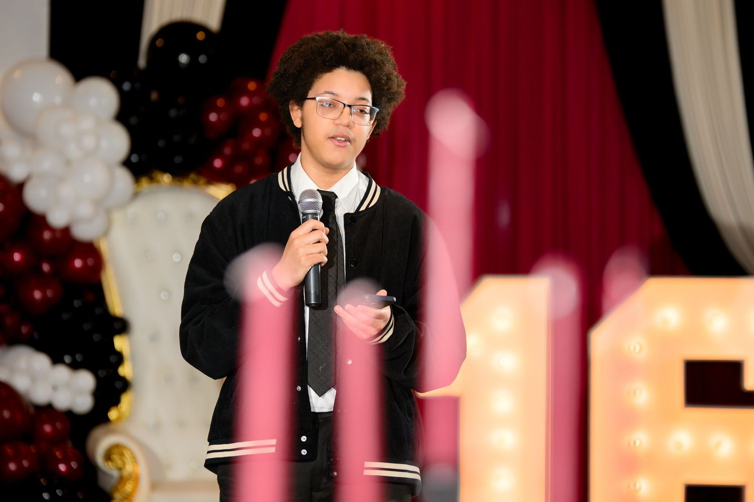 Young man with curly hair speaking into a microphone at an event with balloon decorations.