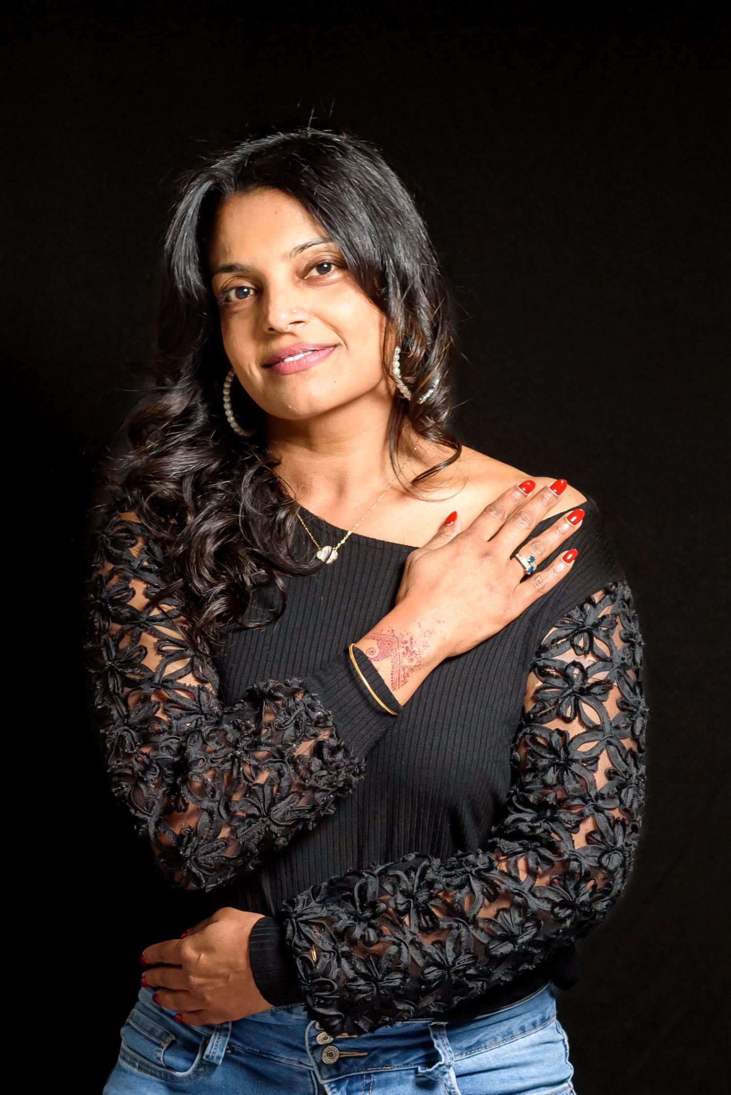 A woman in a black lace top and jeans poses confidently against a dark background, wearing gold earrings and a necklace.