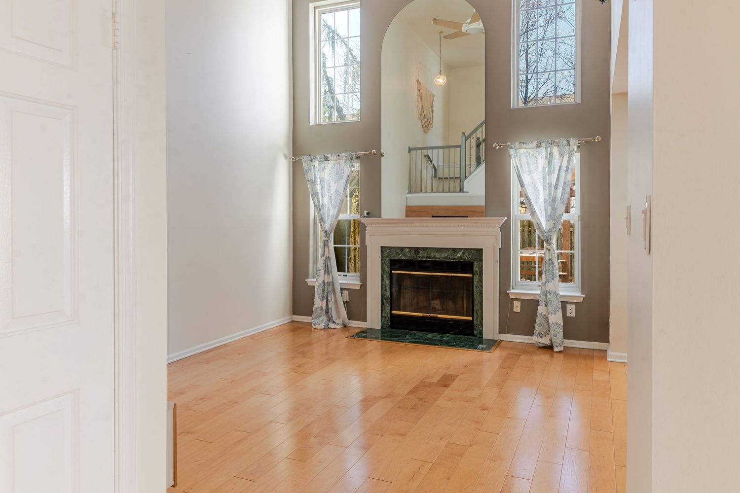 Elegant living room with hardwood floors, electric fireplace, arched mirror, and soaring two-story windows with sheer curtains.