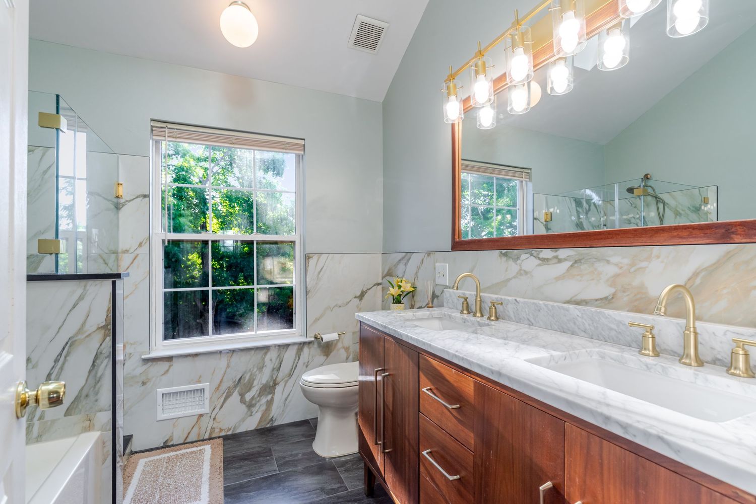 Elegant bathroom with marble walls, wood vanity, gold fixtures, dual sinks, and a walk-in shower with vaulted ceilings.