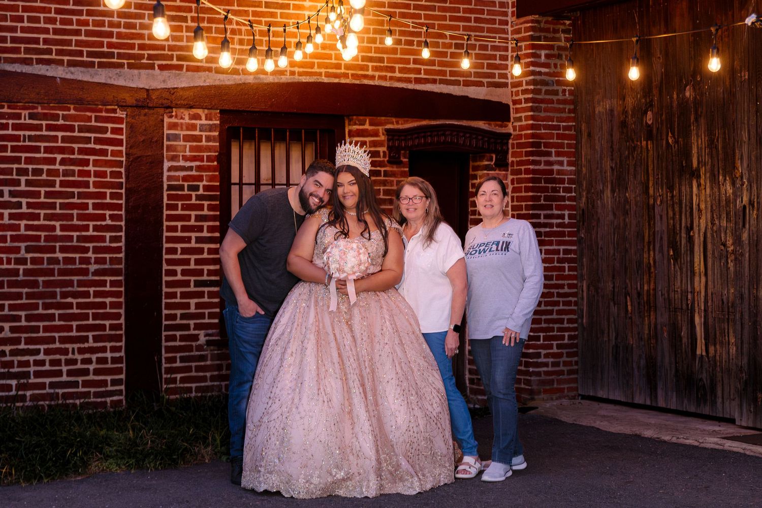 A group stands together in front of a brick wall and wooden door under warm string lights at dusk.