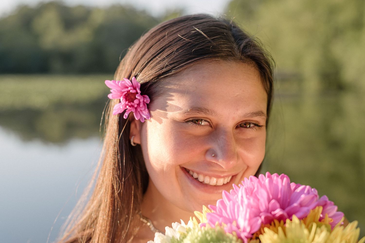 A pink flower adorns long dark hair as someone poses with a bouquet by a serene lake on a sunny day.