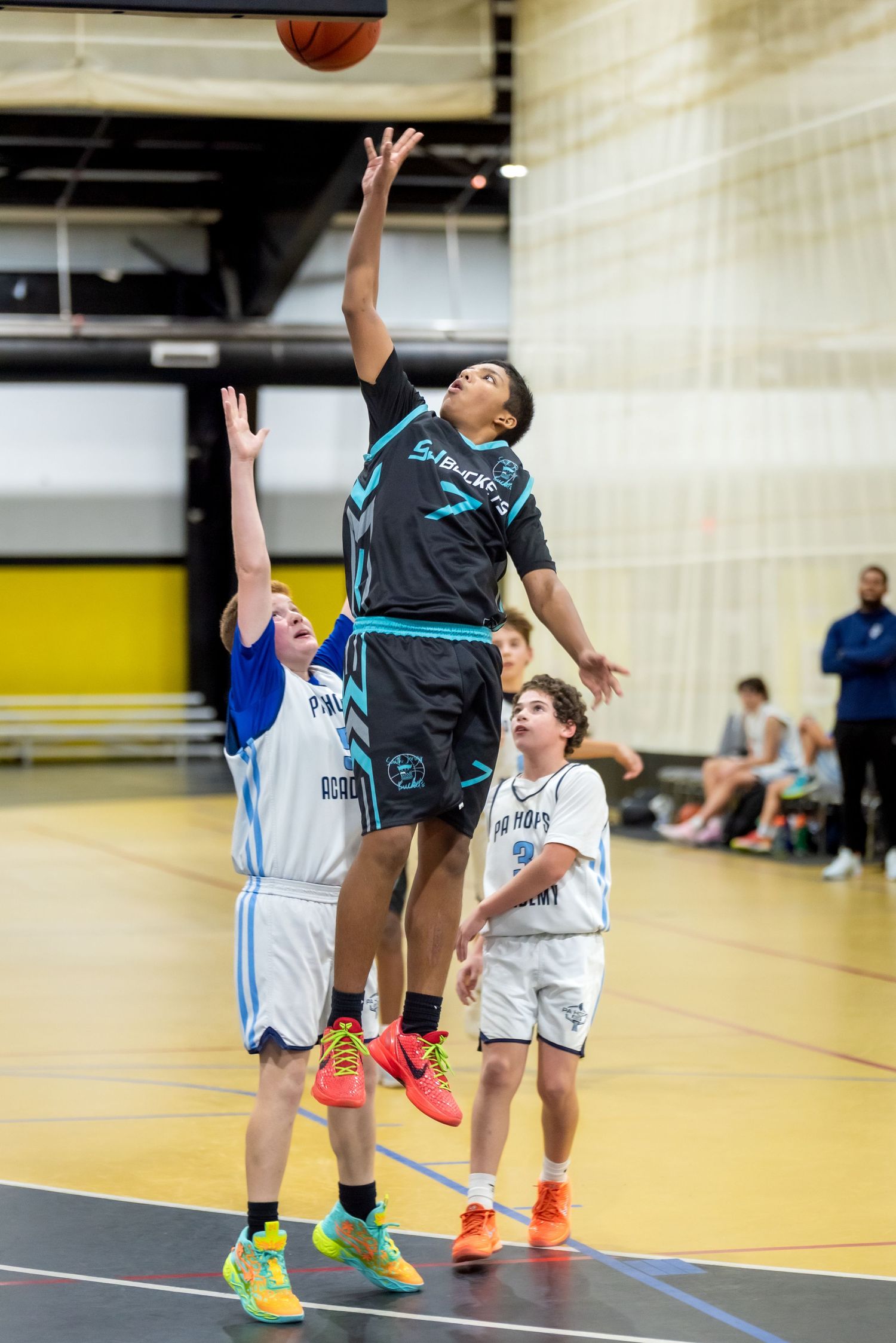 Young basketball player in black uniform leaps high to shoot over defenders during an indoor youth basketball game.