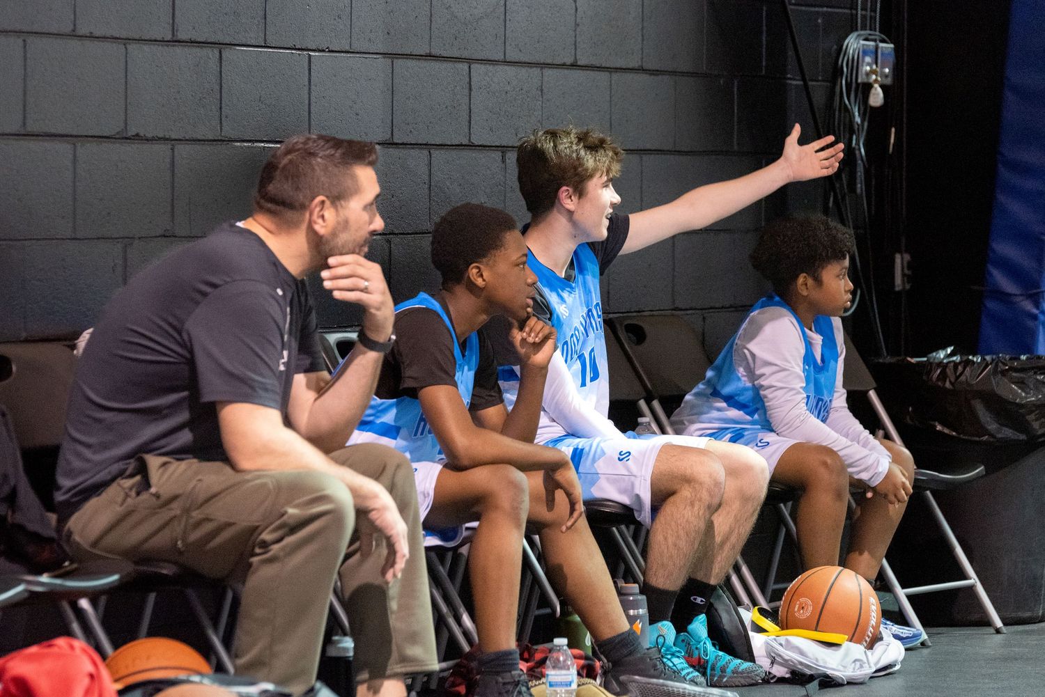 Youth basketball players sit on bench with coach during game timeout at indoor sports facility.