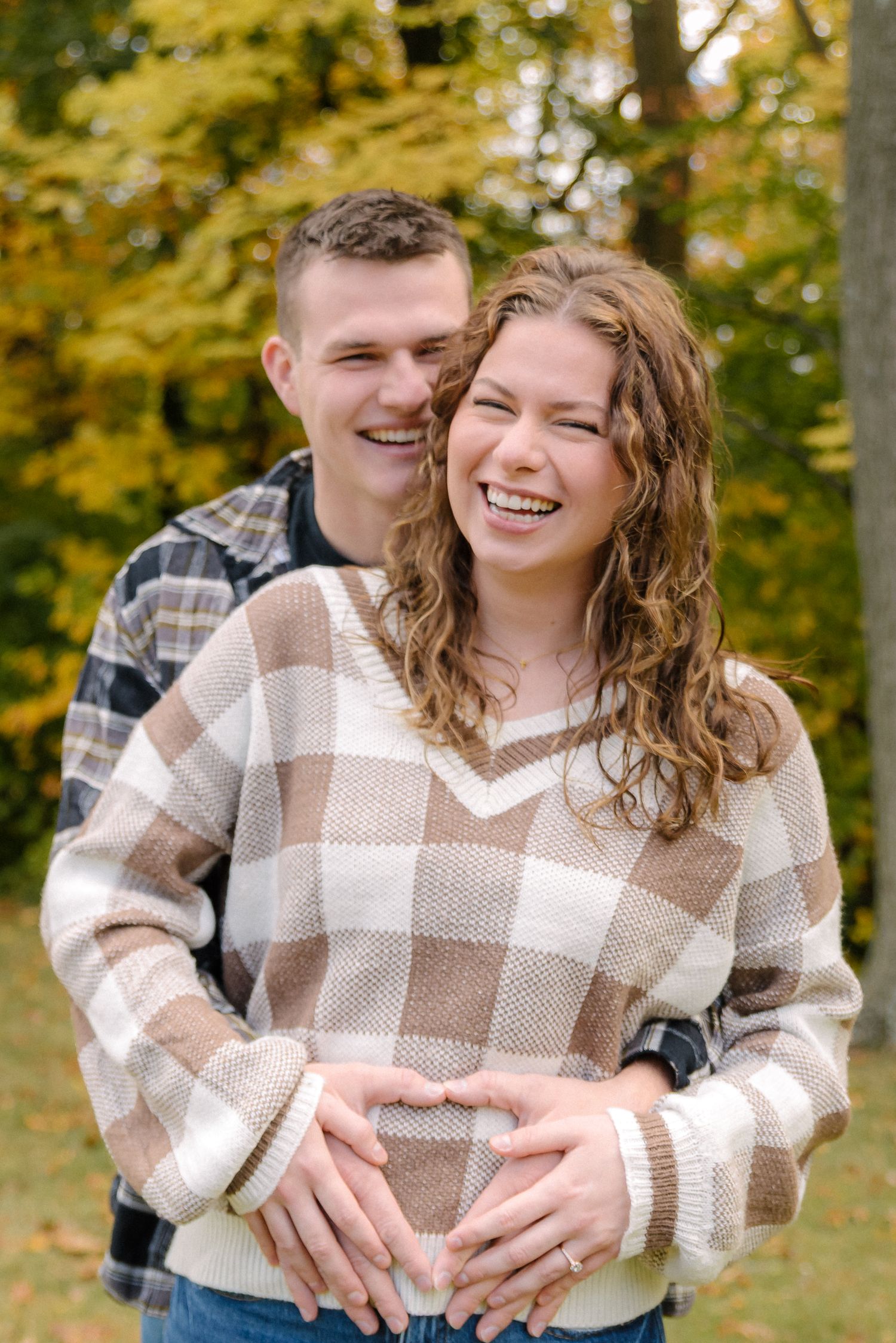A couple wearing matching plaid sweaters share joyful moments together in a fall setting with yellow leaves in background.