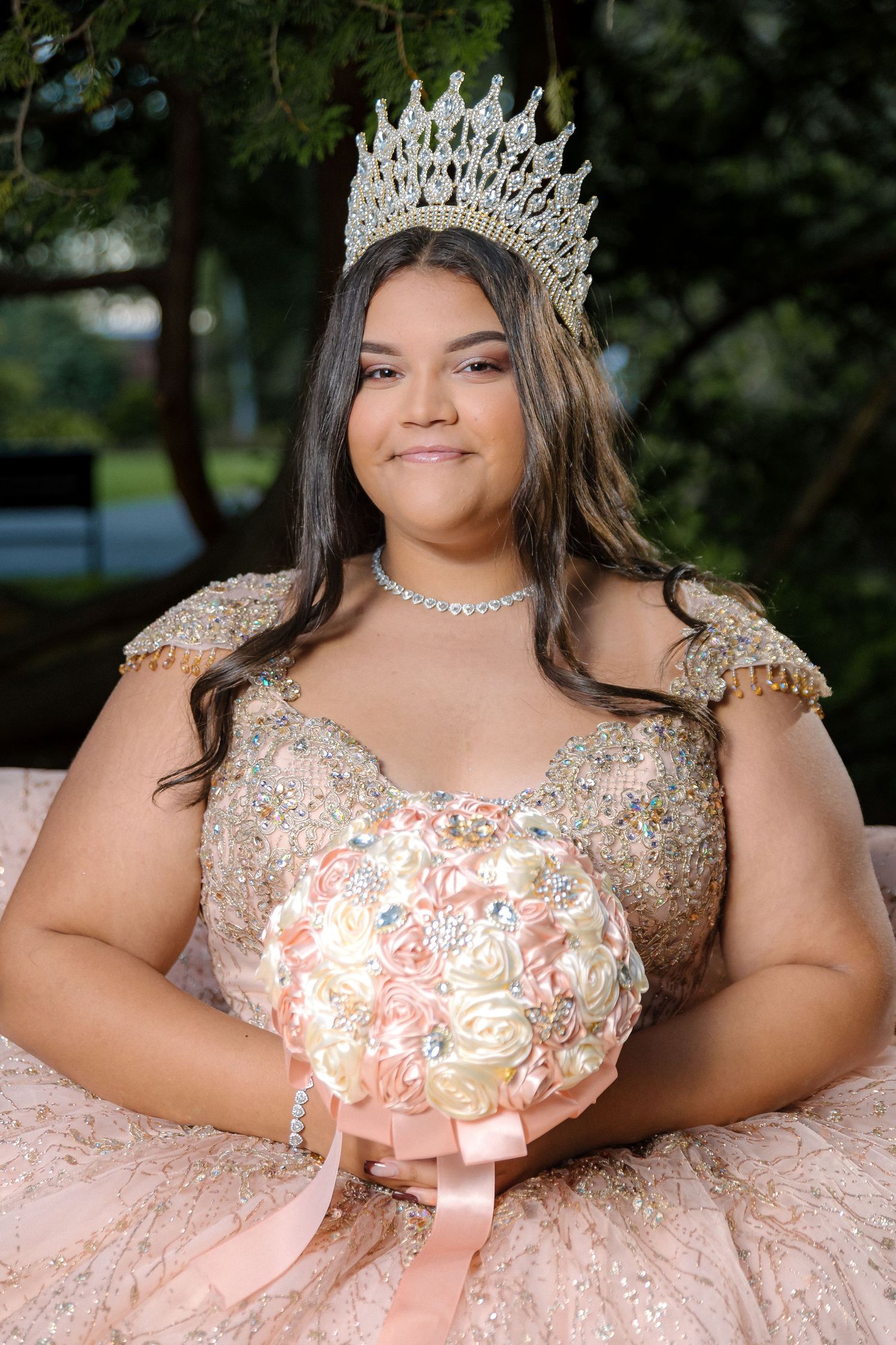 A person wearing a sparkling pink ballgown and crystal crown poses outdoors in a garden setting.