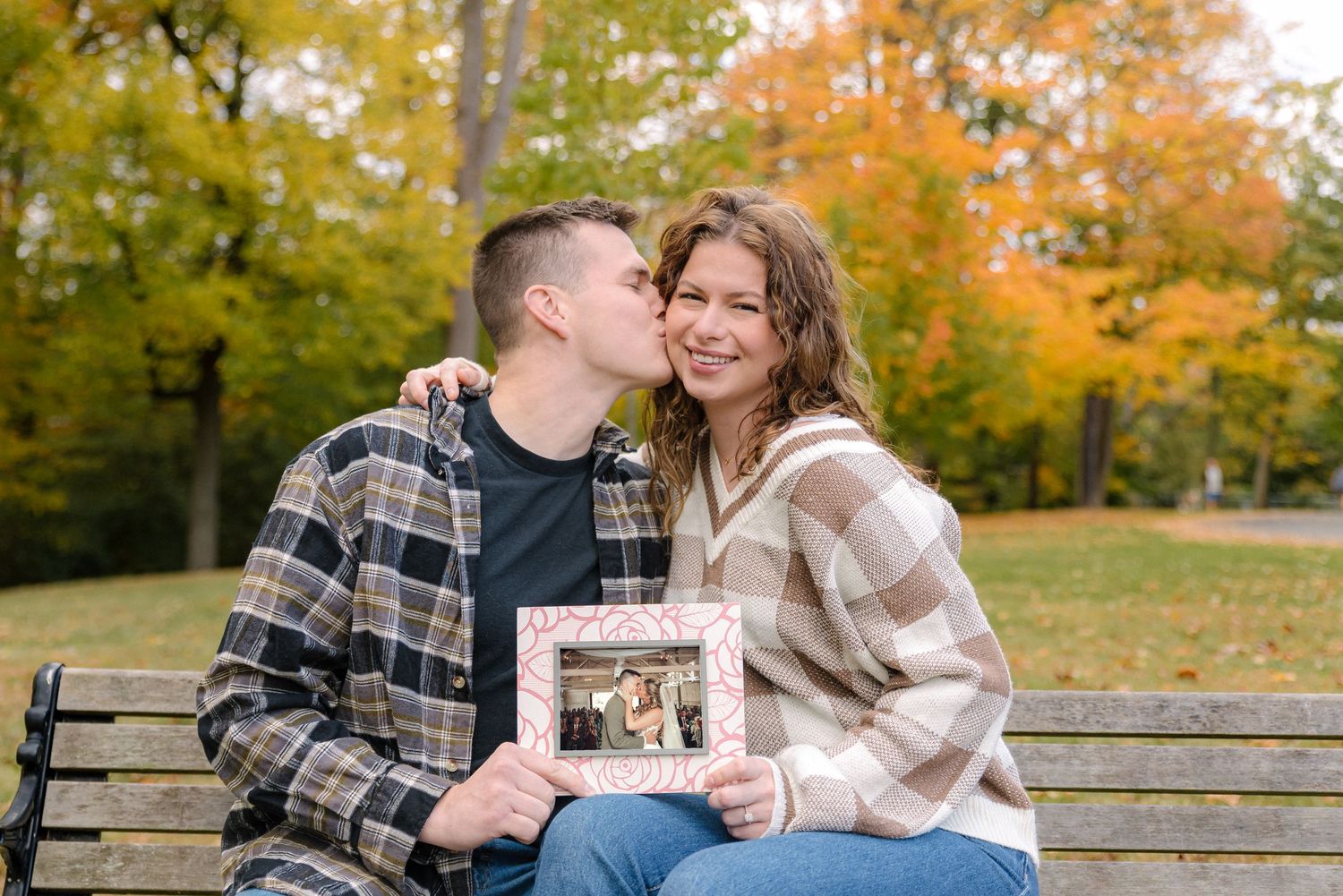 A couple sits together on a wooden bench surrounded by vibrant fall foliage in a park setting.