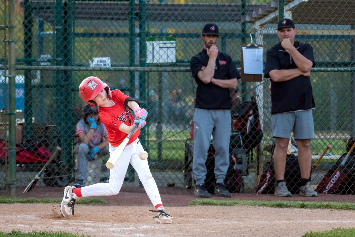 Young baseball player in red uniform swings bat during little league game with coaches watching in background.