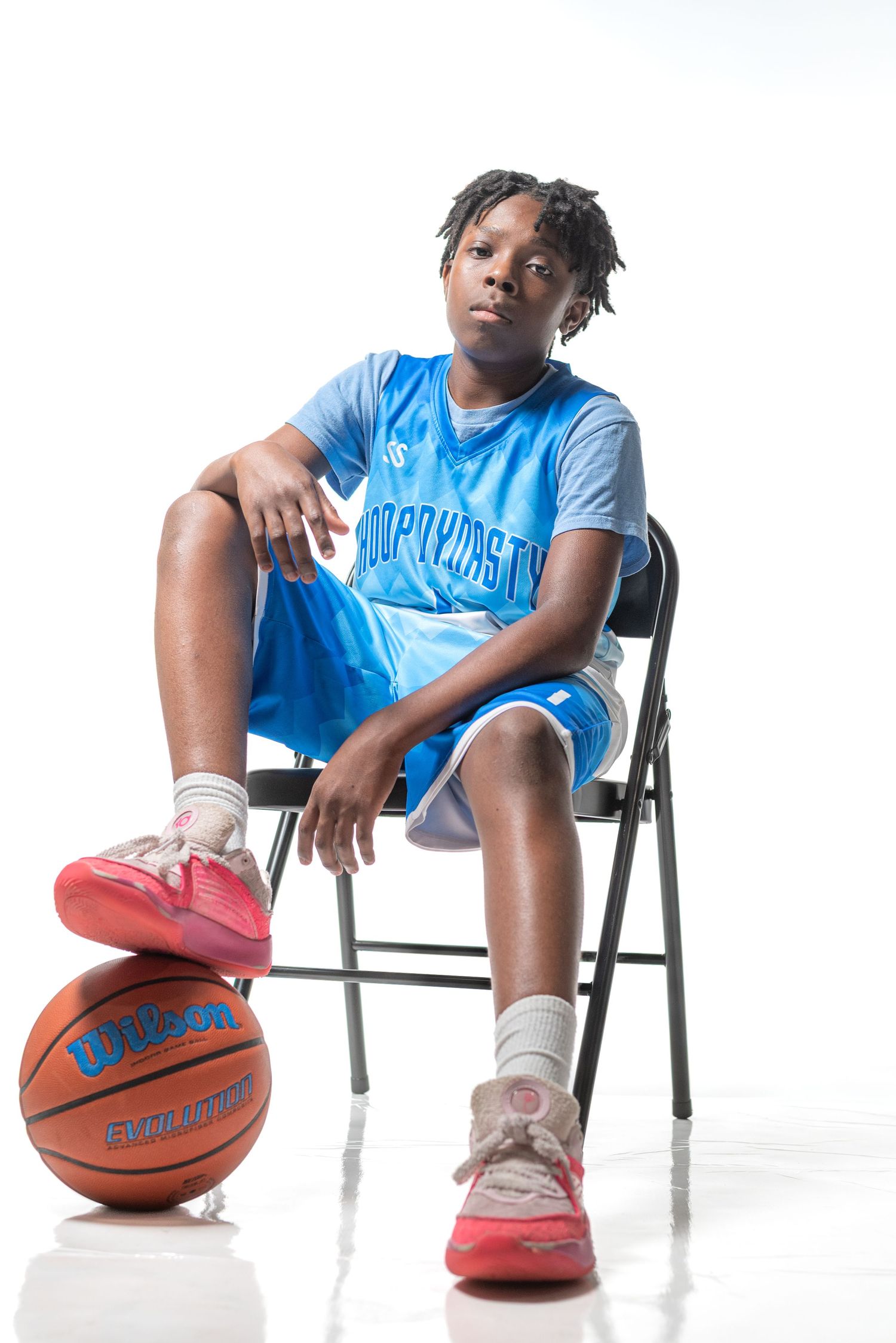 Young basketball player in blue uniform sitting on folding chair with Wilson basketball, white studio background.