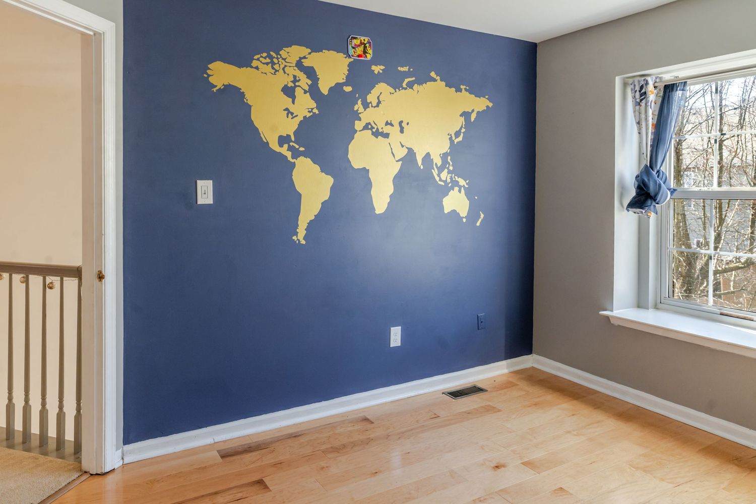 Bedroom with blue accent wall featuring gold world map decal, hardwood floors, and natural light from window.