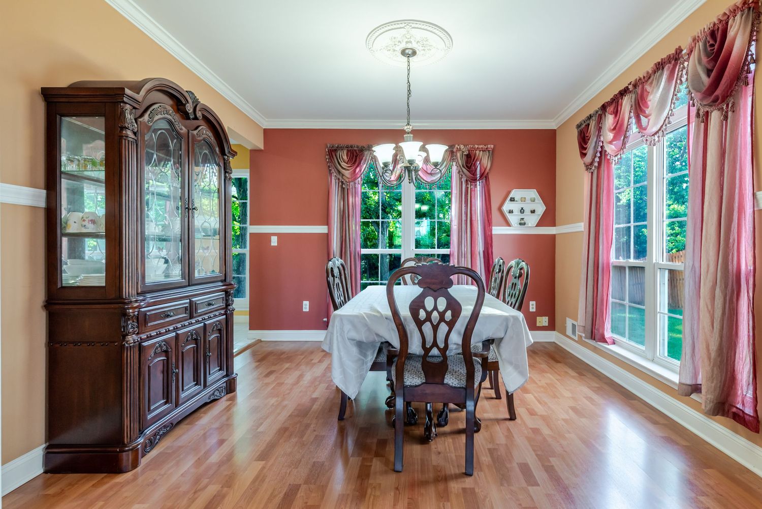 Elegant dining room with hardwood floors, cherry wood china cabinet, chandelier, and large windows with pink curtains.