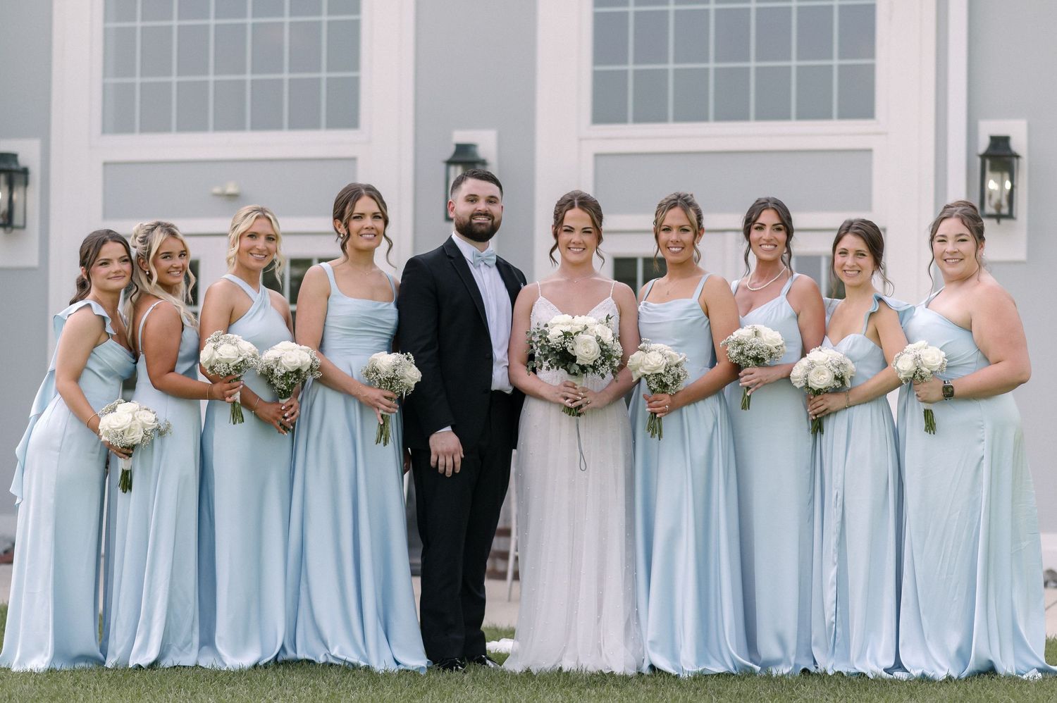 Groom in black tuxedo poses with bride in white gown and eight bridesmaids in light blue dresses holding white bouquets.