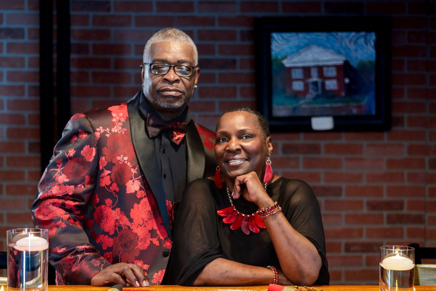Couple posing together, man in red floral blazer and woman in black outfit with red necklace, brick wall background.